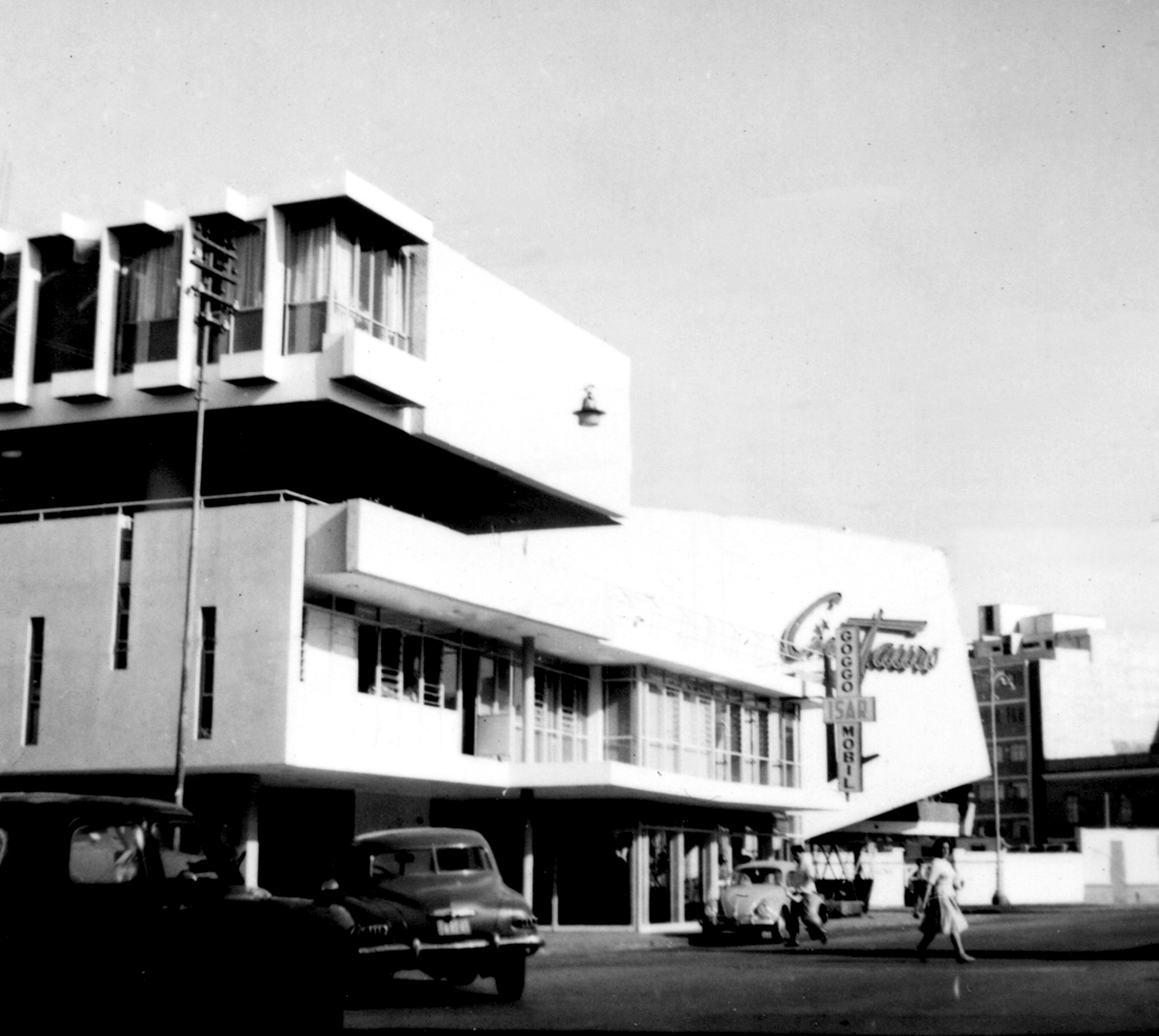 Angled facade showing the projecting canopy and street-level entrance with vintage cars passing by