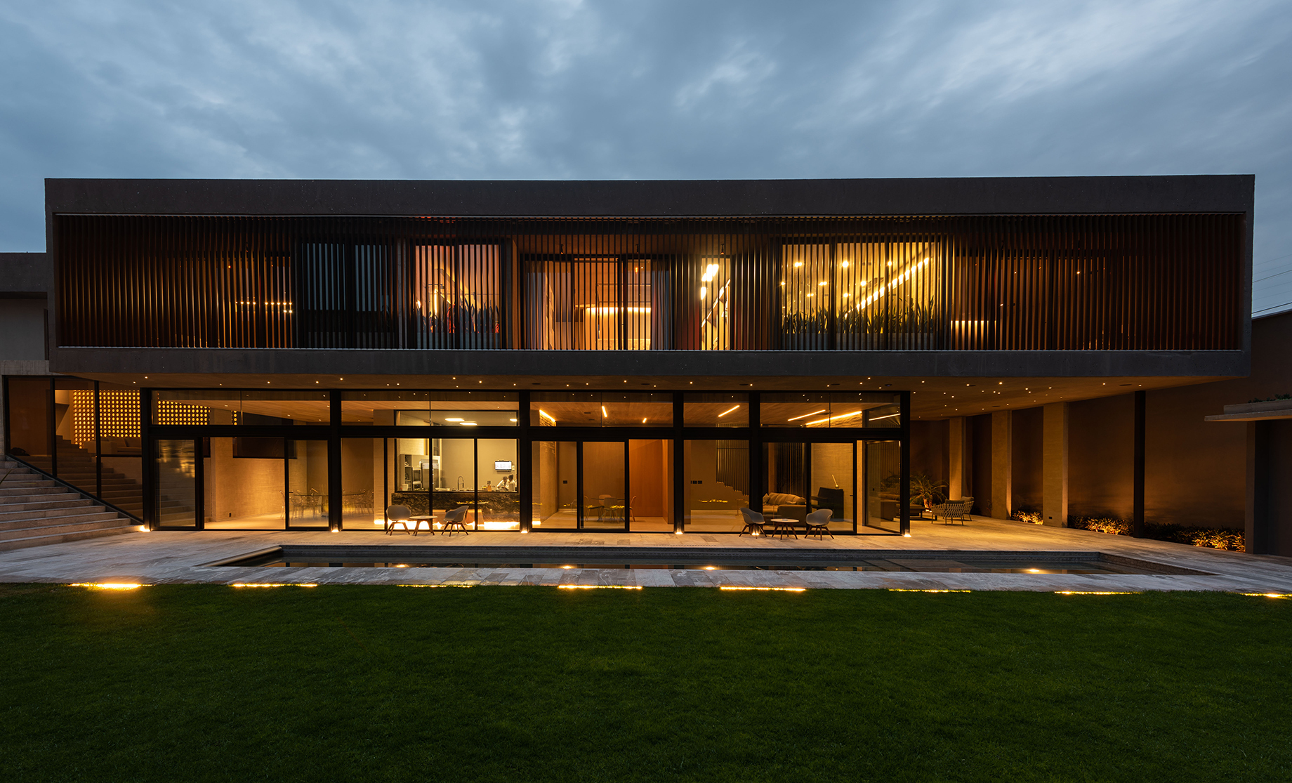 Two-storey concrete volume with timber slat screen above a glazed ground floor, lit warmly at dusk beside a lawn and pool edge