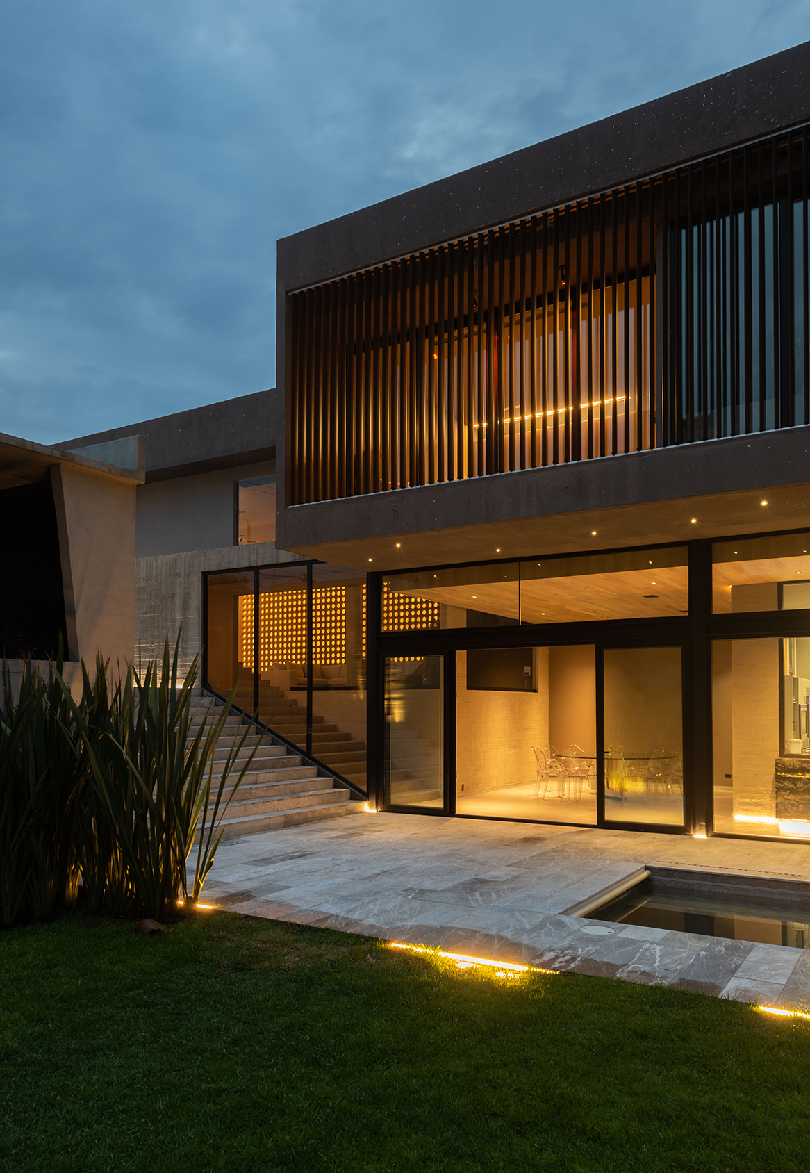 Corner of the slatted upper volume above glazed living space, with stone steps and agave plants under a dark blue evening sky