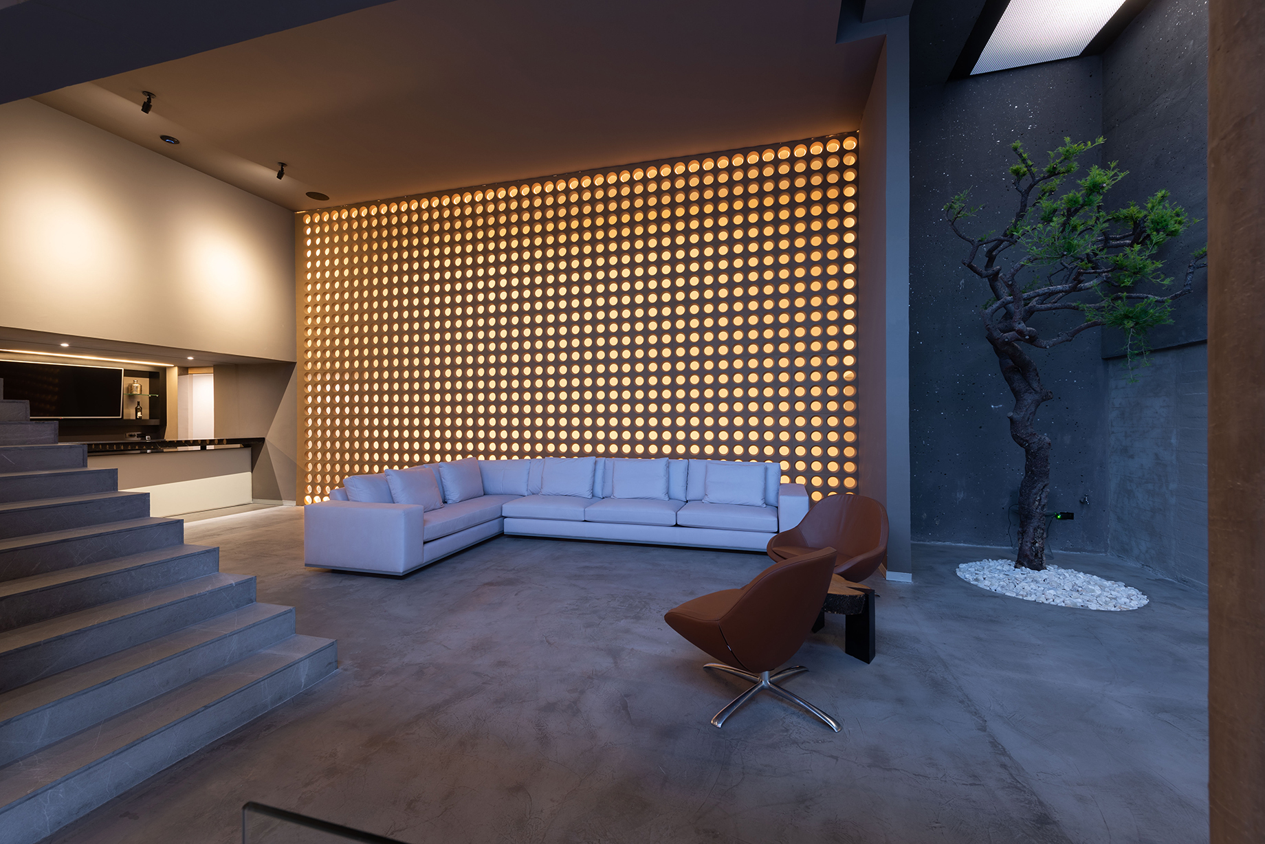 Lounge with a white corner sofa before a backlit perforated wall, a sculpted tree in a pebble bed beside it