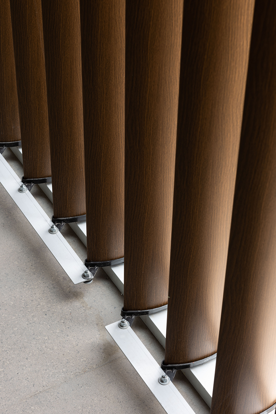 Close-up of round wood-finish vertical slats fixed to white metal floor tracks with steel brackets and bolts