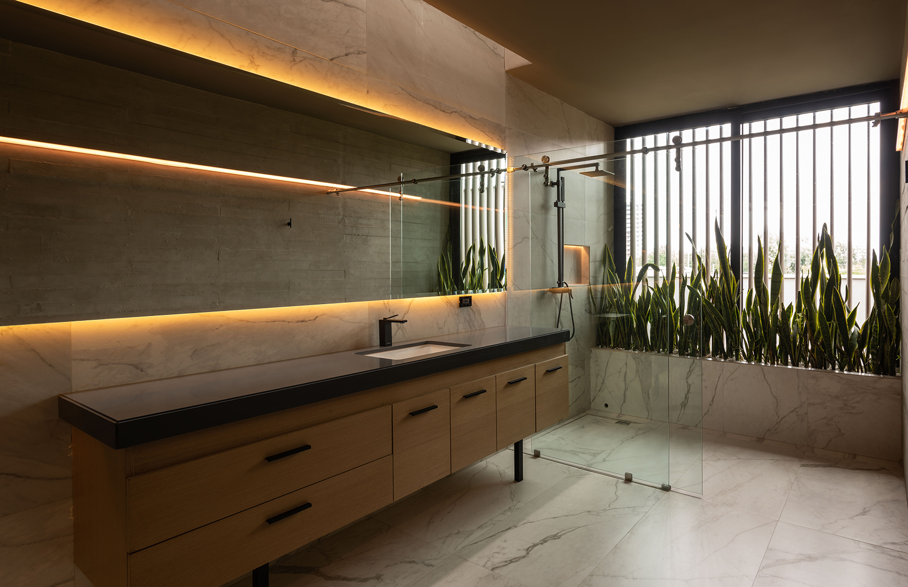 Marble bathroom with long timber vanity, backlit mirror and a glass shower beside a planter of snake plants in daylight