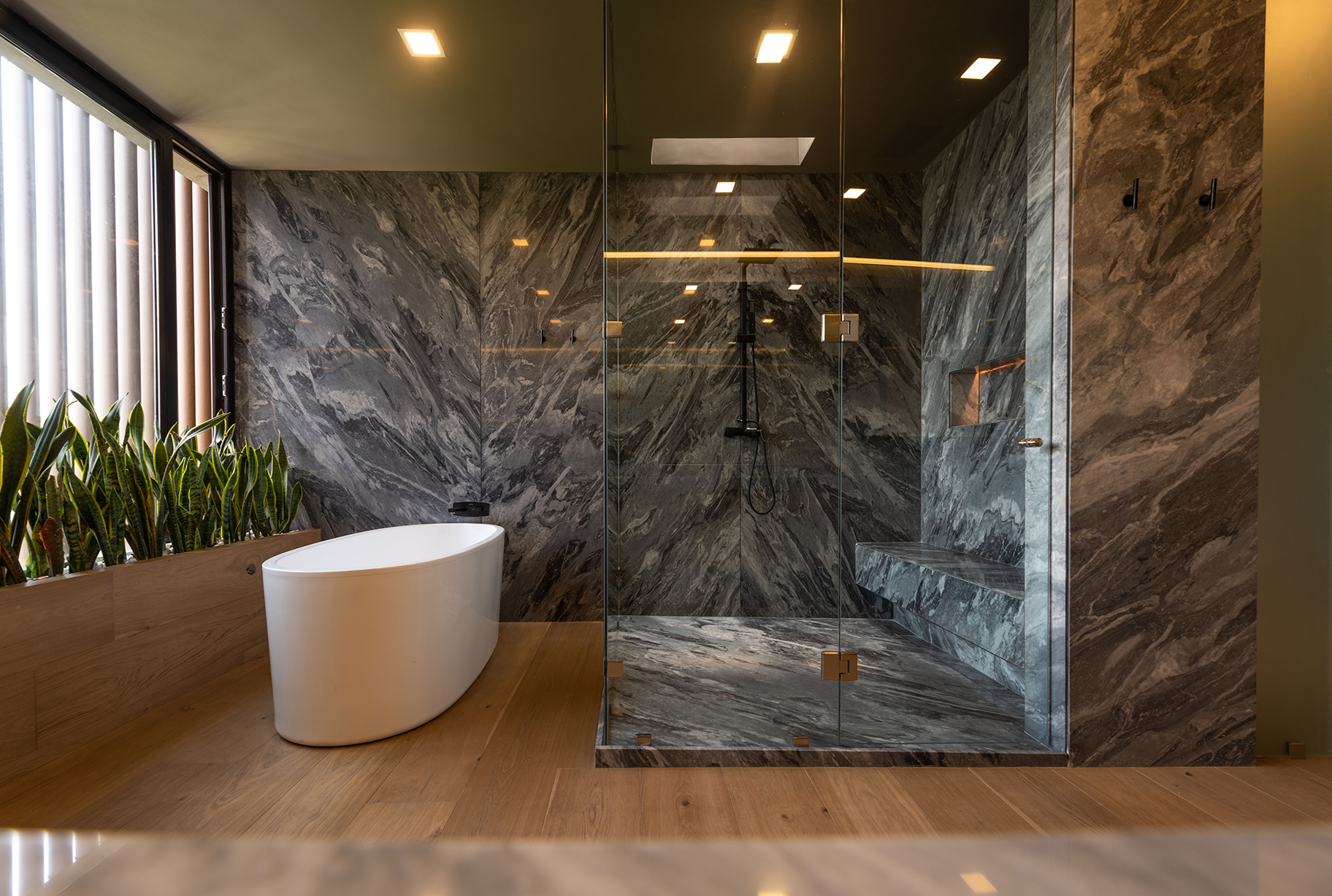 Freestanding white tub beside a glass-enclosed shower clad in dark veined stone, with a planter of snake plants by the window