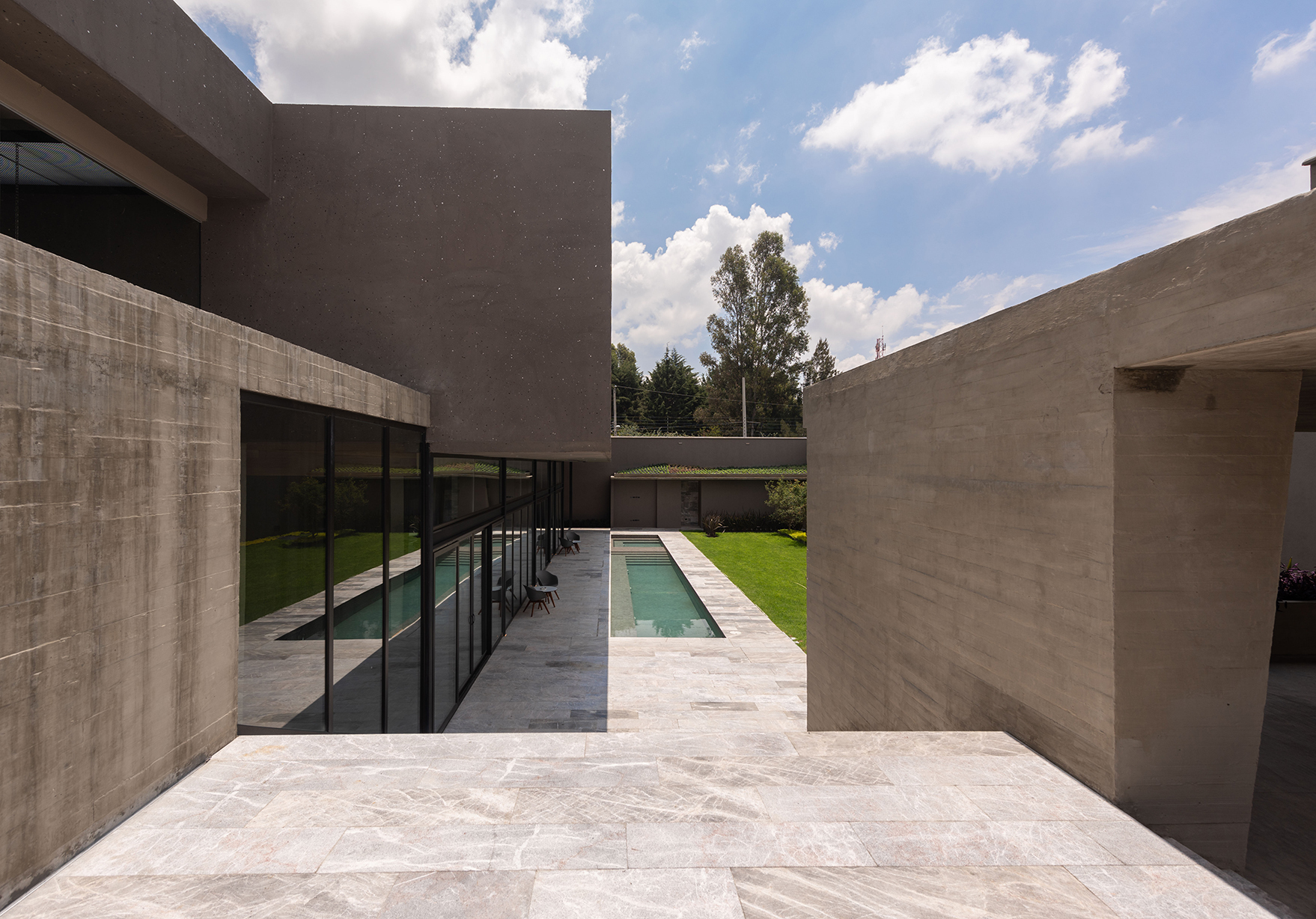 Board-formed concrete walls frame a narrow lap pool and lawn beyond a pale stone terrace under scattered clouds