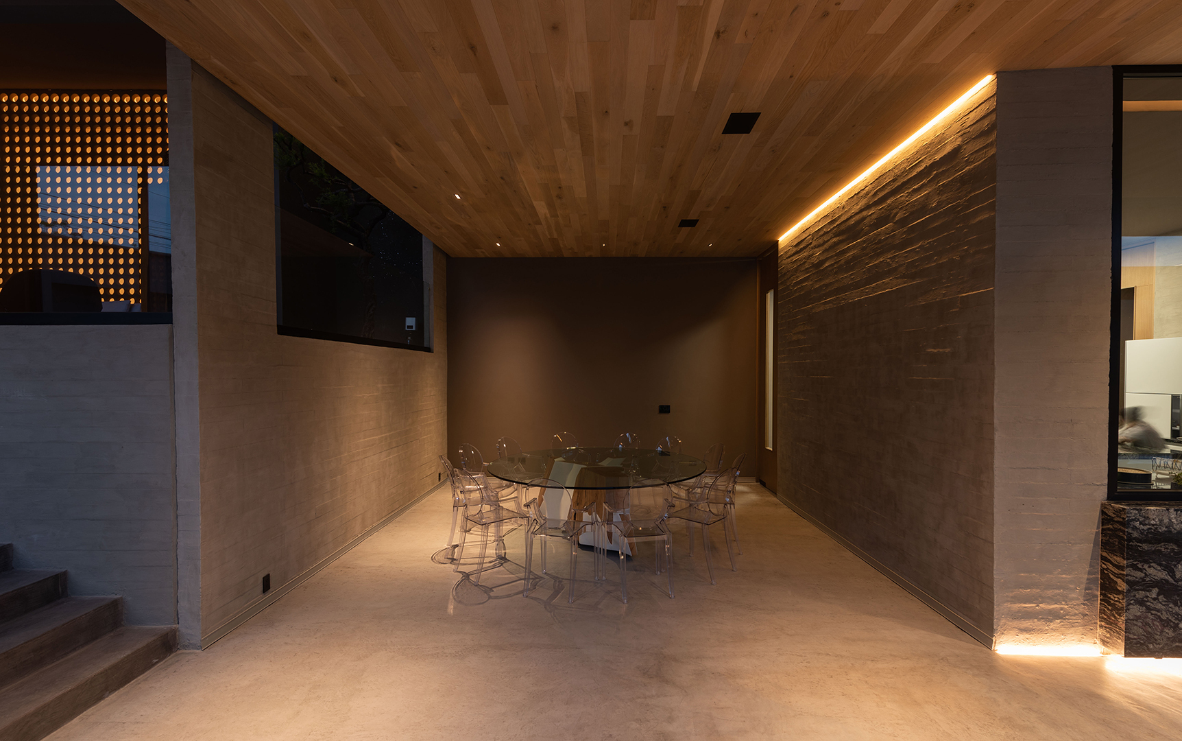 Covered dining area with a round glass table, timber ceiling and concrete wall washed by a linear light at dusk