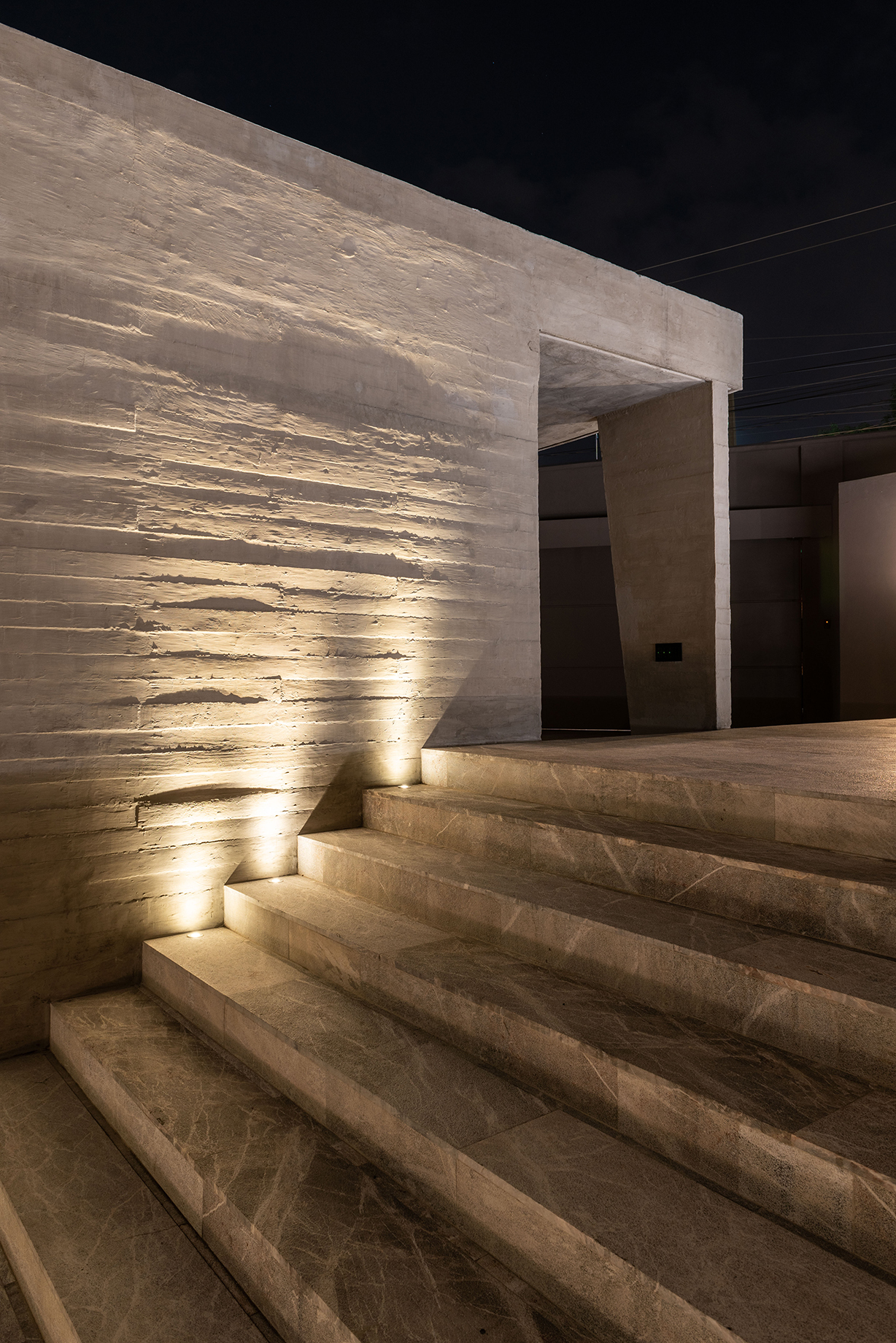 Pale stone steps rising beside a board-formed concrete wall, lit by low ground lights against the night sky