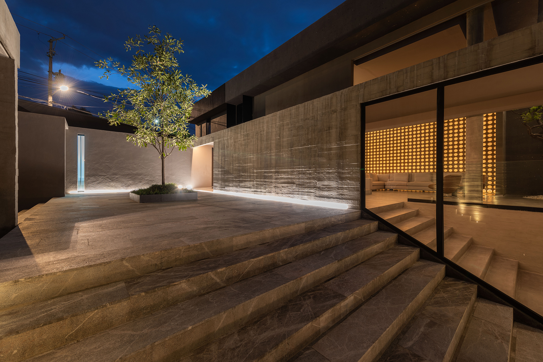 Stone steps lead to a courtyard with a single tree, while a glazed wall reveals the glowing screen inside at twilight