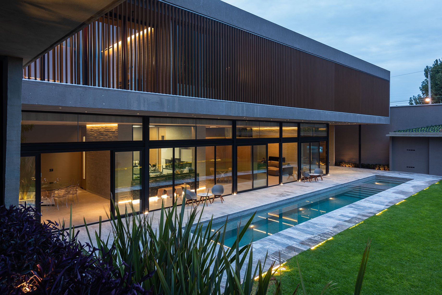 Dusk view of the timber-slatted upper volume above a glazed ground floor, with a lit lap pool and lawn in front