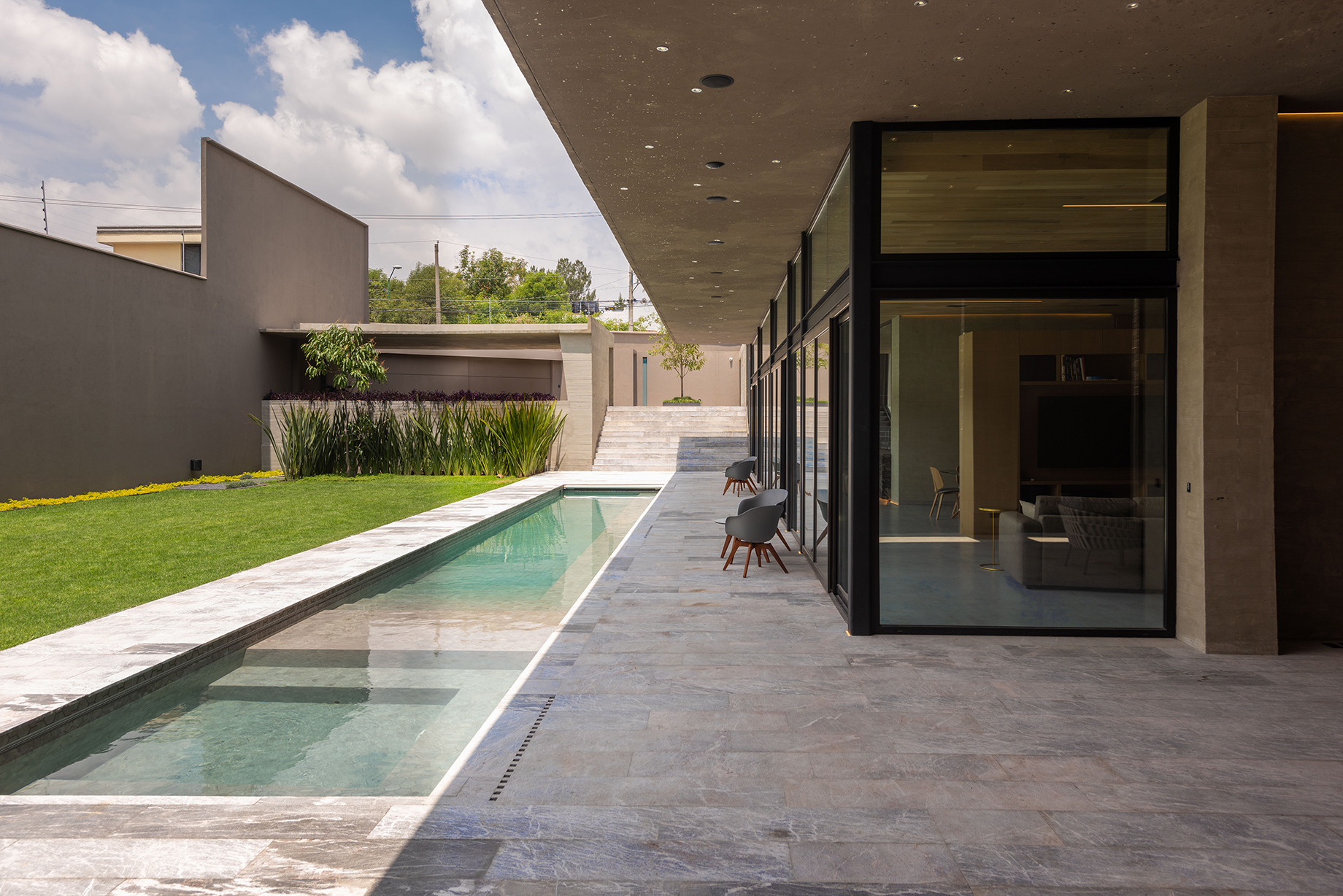 Covered terrace of grey stone paving beside a narrow pool and glass wall, under a partly cloudy sky