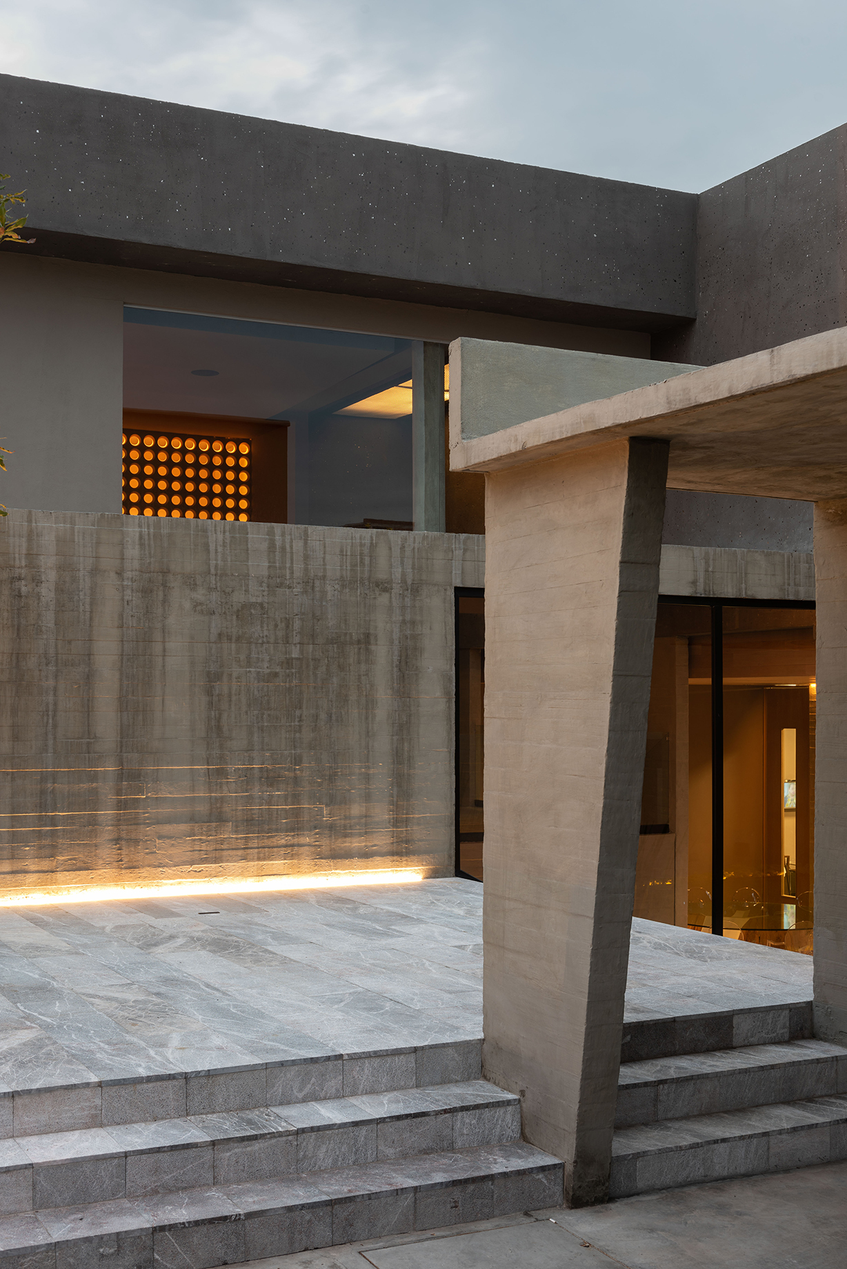 Entry steps of grey stone leading to a slanted concrete column and board-marked wall, lit by a low strip light at dusk