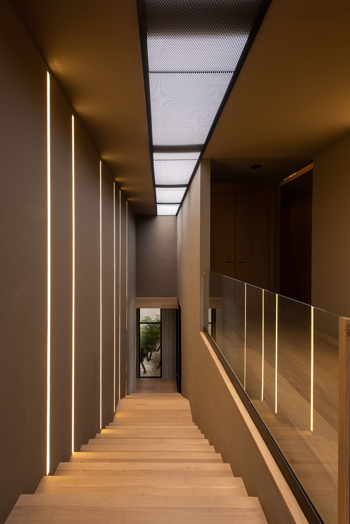 Timber staircase lined with recessed vertical light strips, a glass balustrade and a perforated metal skylight overhead