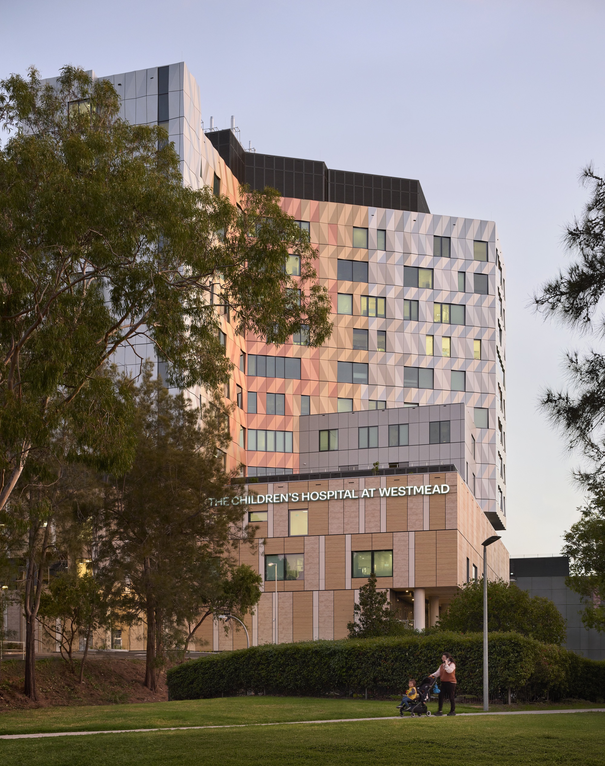 Tower facade with triangular panel cladding in warm and cool tones beside a lawn at twilight