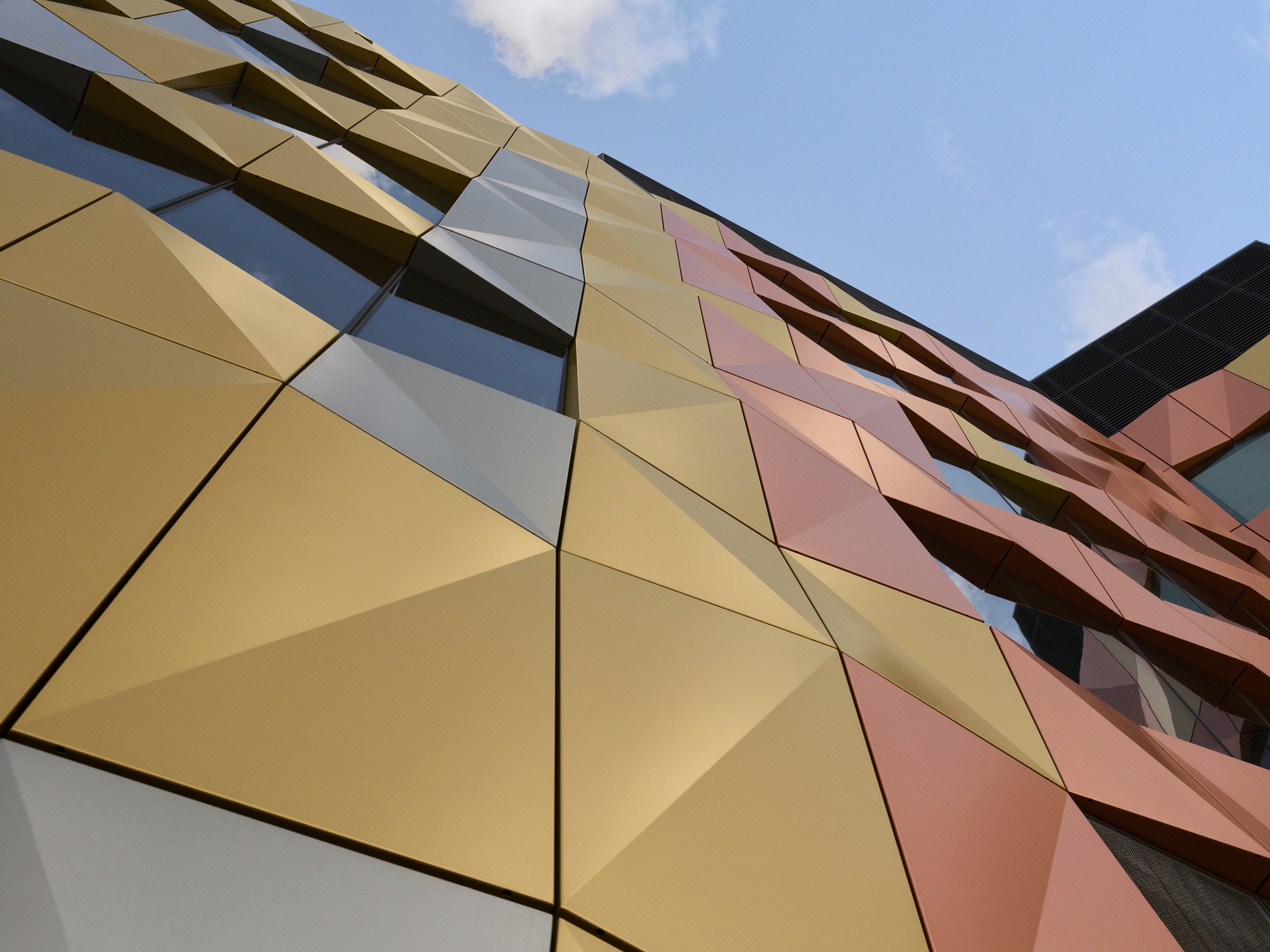 Upward view of the geometric metal panel facade in gold, pink, and copper tones against a blue sky
