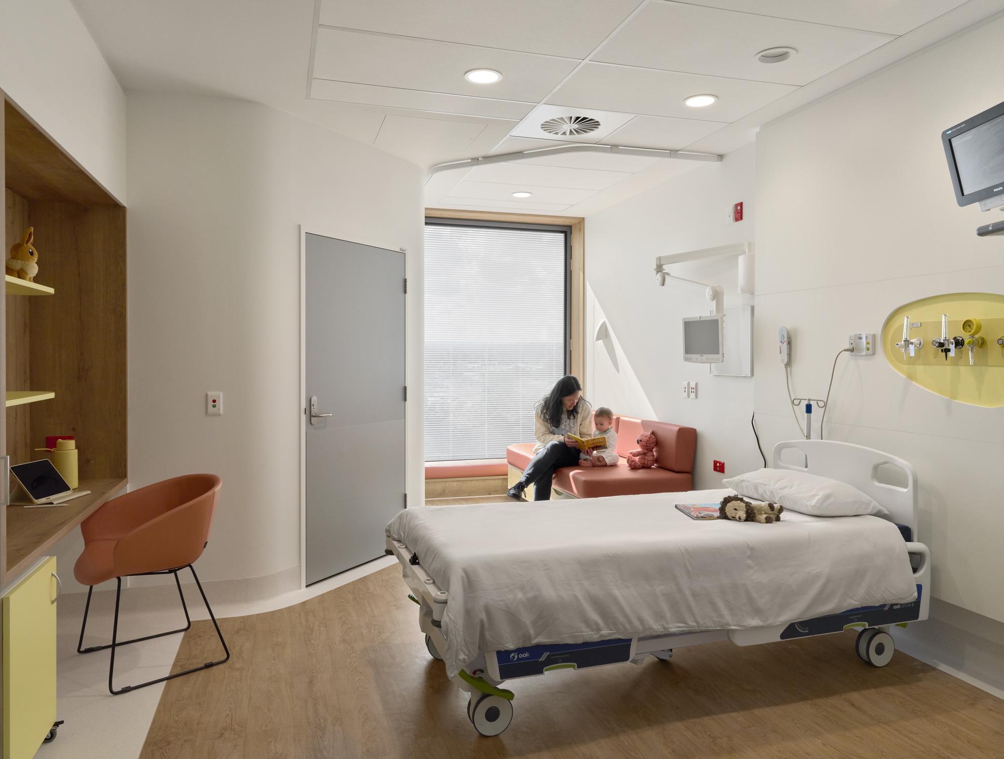 Patient room with hospital bed, window seat, and wooden desk under a suspended ceiling with recessed lighting