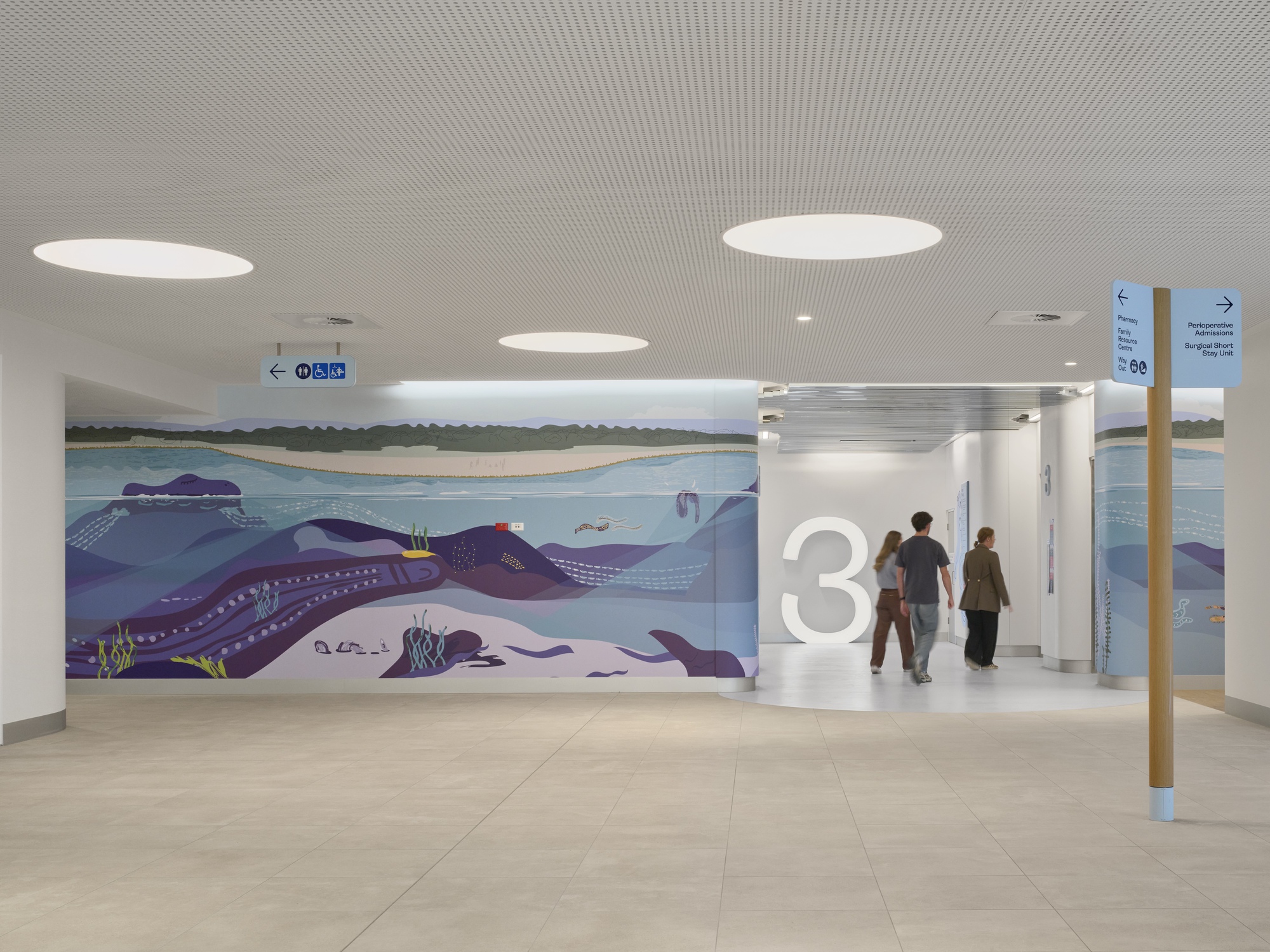 Hospital corridor with painted underwater mural featuring octopi and large circular skylights in perforated metal ceiling