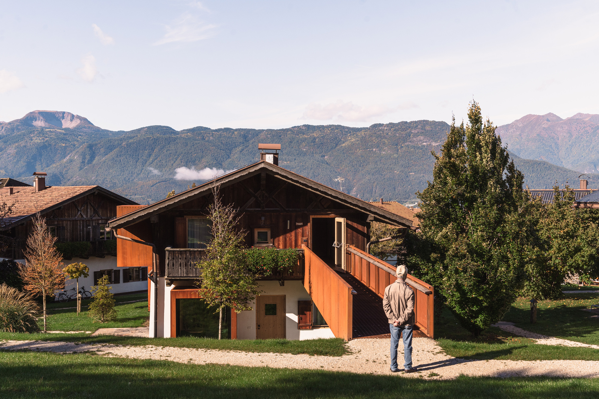Timber chalet with orange metal stair and balcony panels beneath mountain peaks with a visitor walking