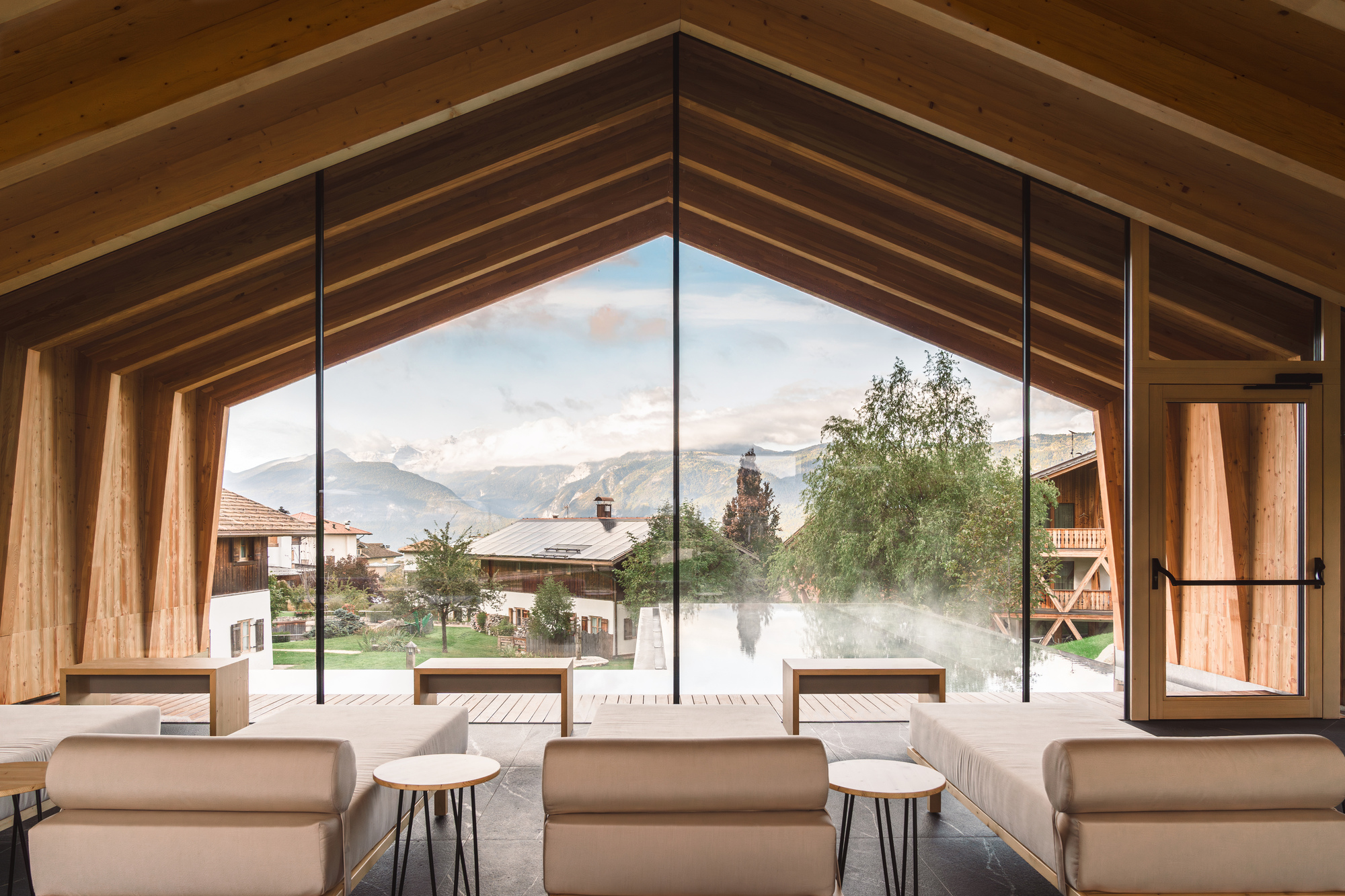 Lounge space beneath timber beam ceiling framing a view through glazing to mist-covered pool terrace