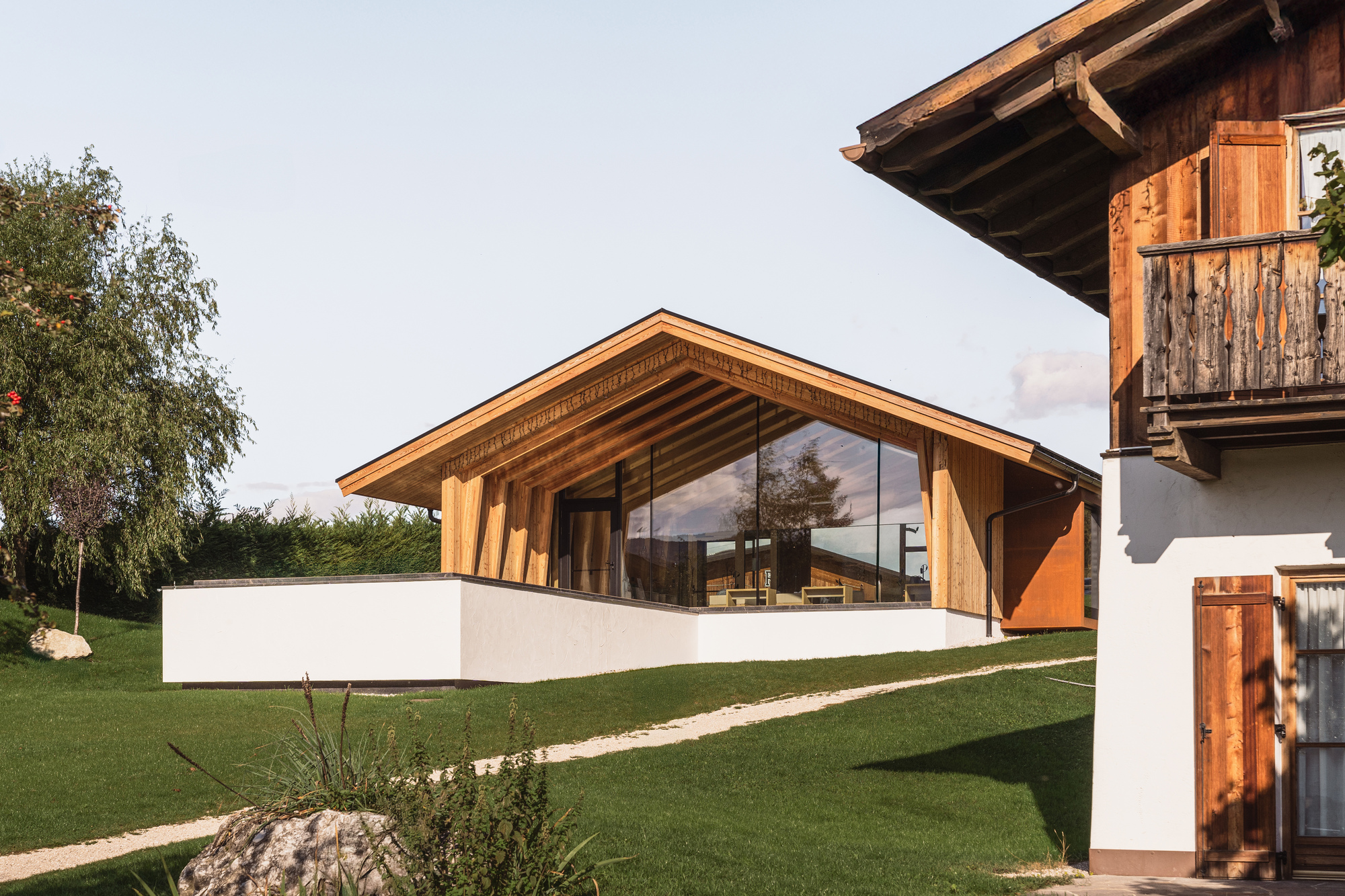 Gabled timber and glass pavilion with white plinth base on a sloped lawn beside existing chalet
