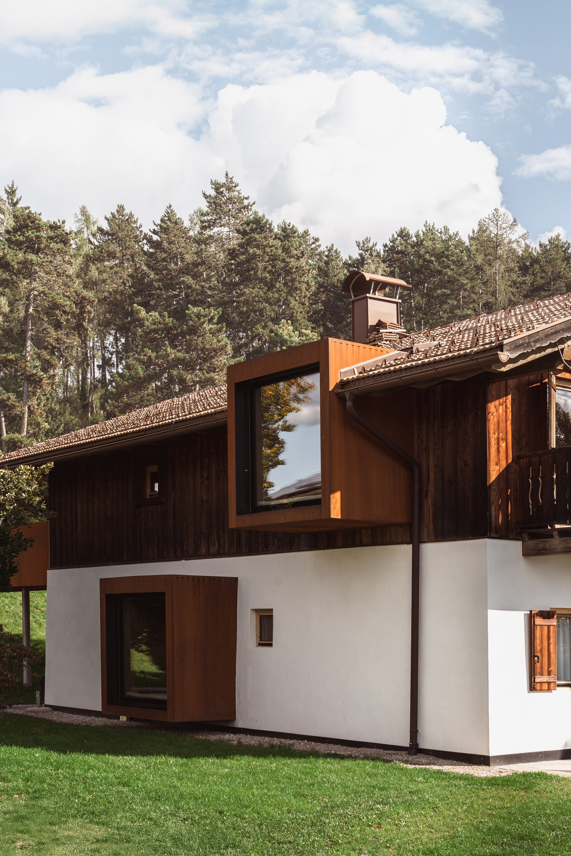 White stucco base with weathered steel bay windows projecting from dark timber upper floor under cloudy sky