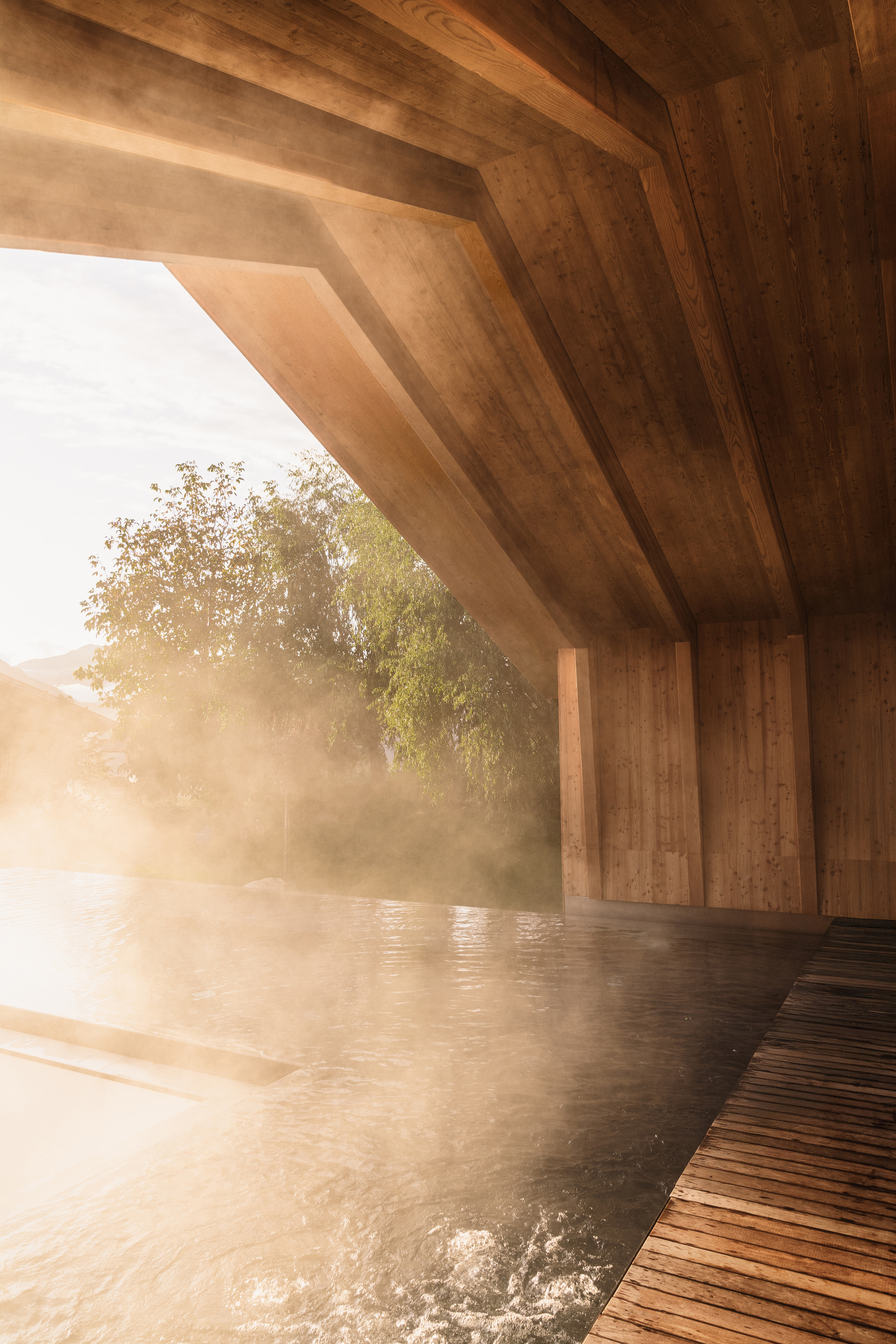 Cantilevered timber roof over steaming pool with angled wood beams in morning mist