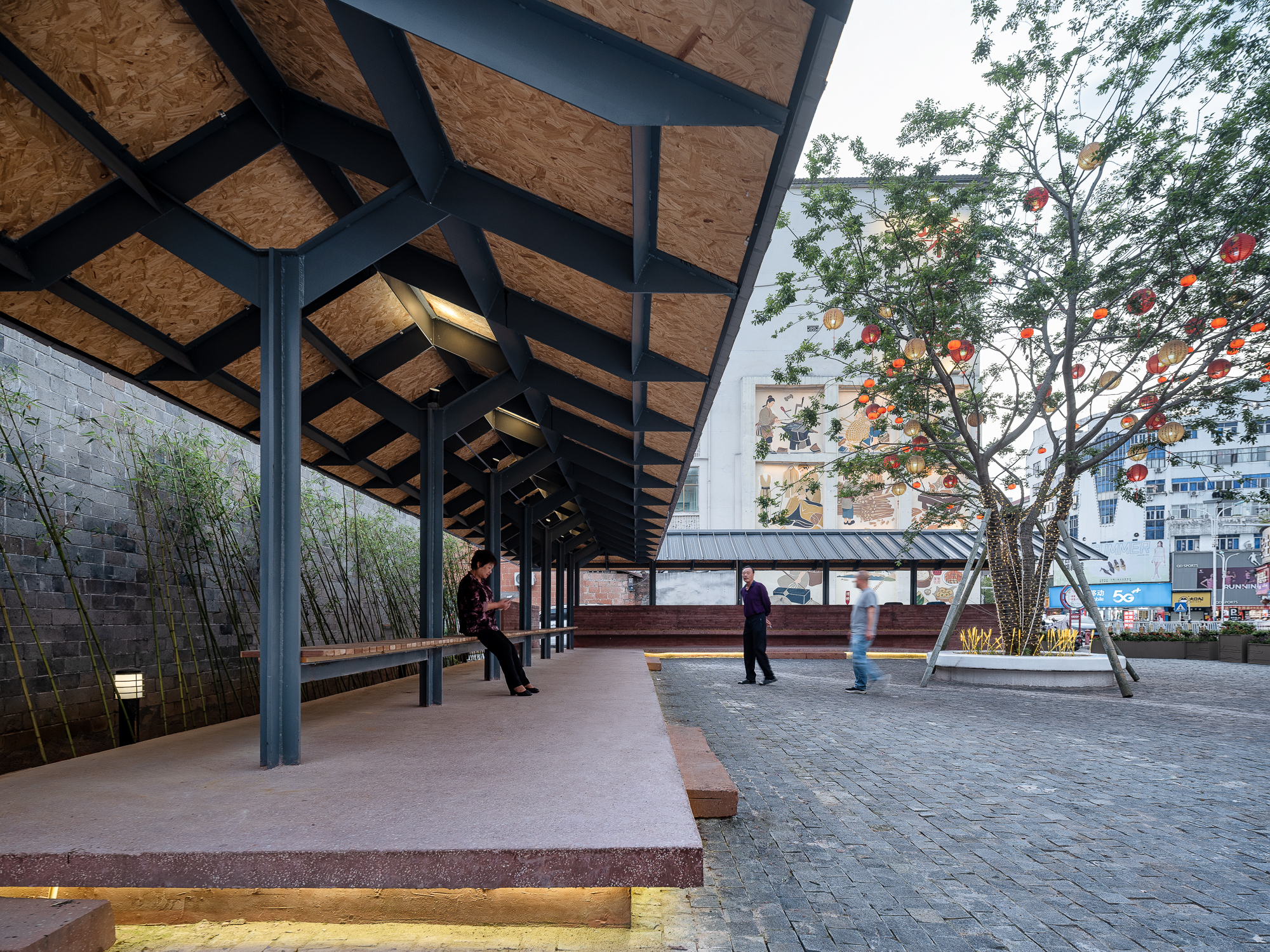 Covered pavilion with black steel columns and oriented strand board ceiling panels in a public plaza with lantern-lit trees