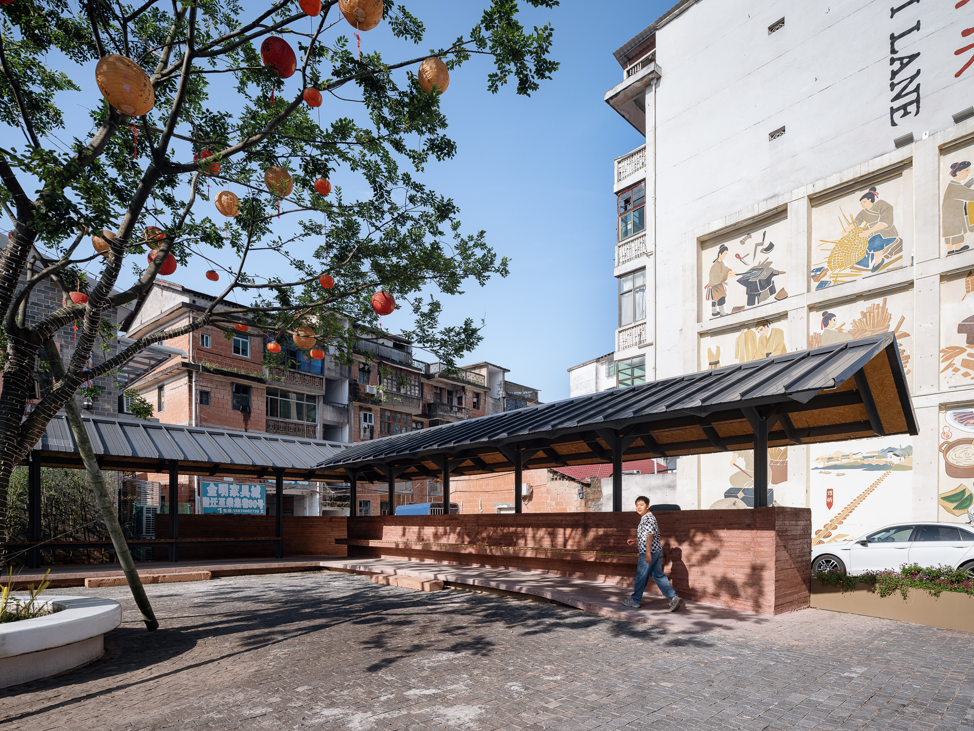 Sheltered courtyard structure with red brick base and metal roof beneath a tree decorated with colored lanterns