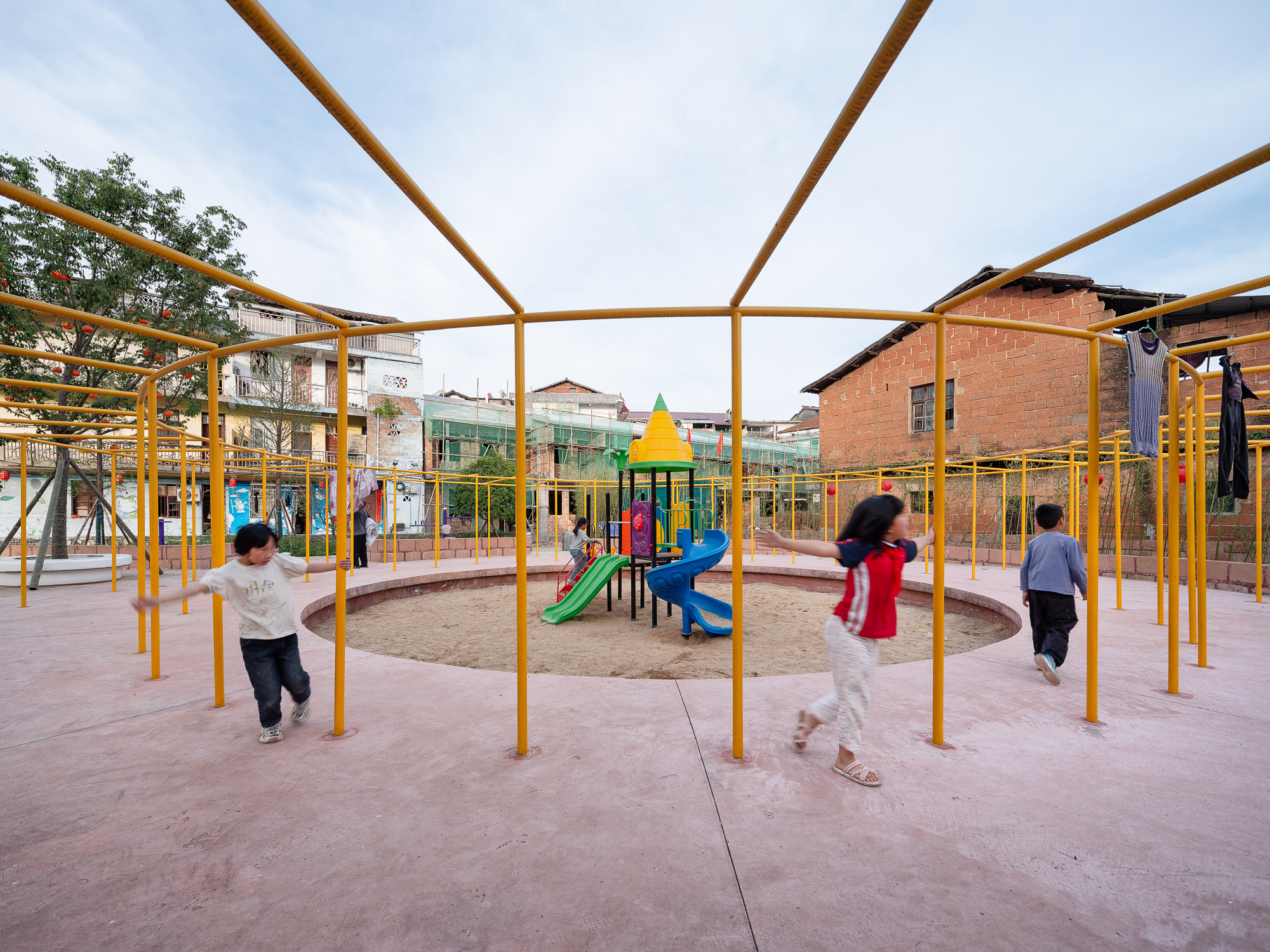 Yellow steel tube canopy sheltering a playground with children running beneath at dusk