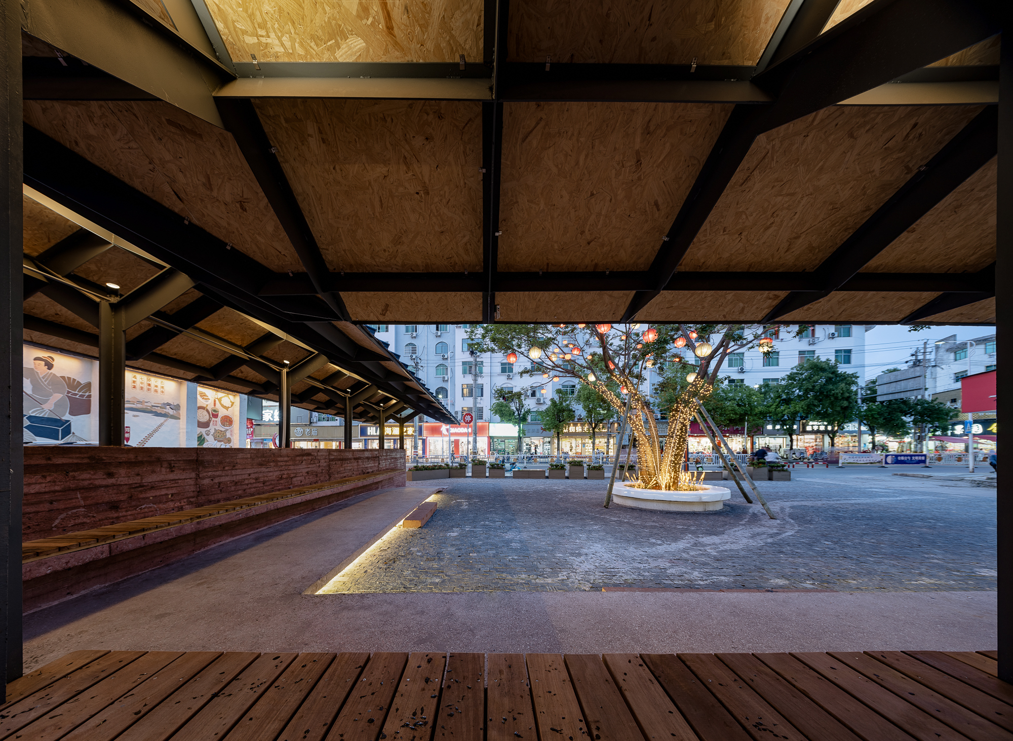 Covered timber deck with plywood ceiling panels and black steel beams overlooking a plaza at twilight