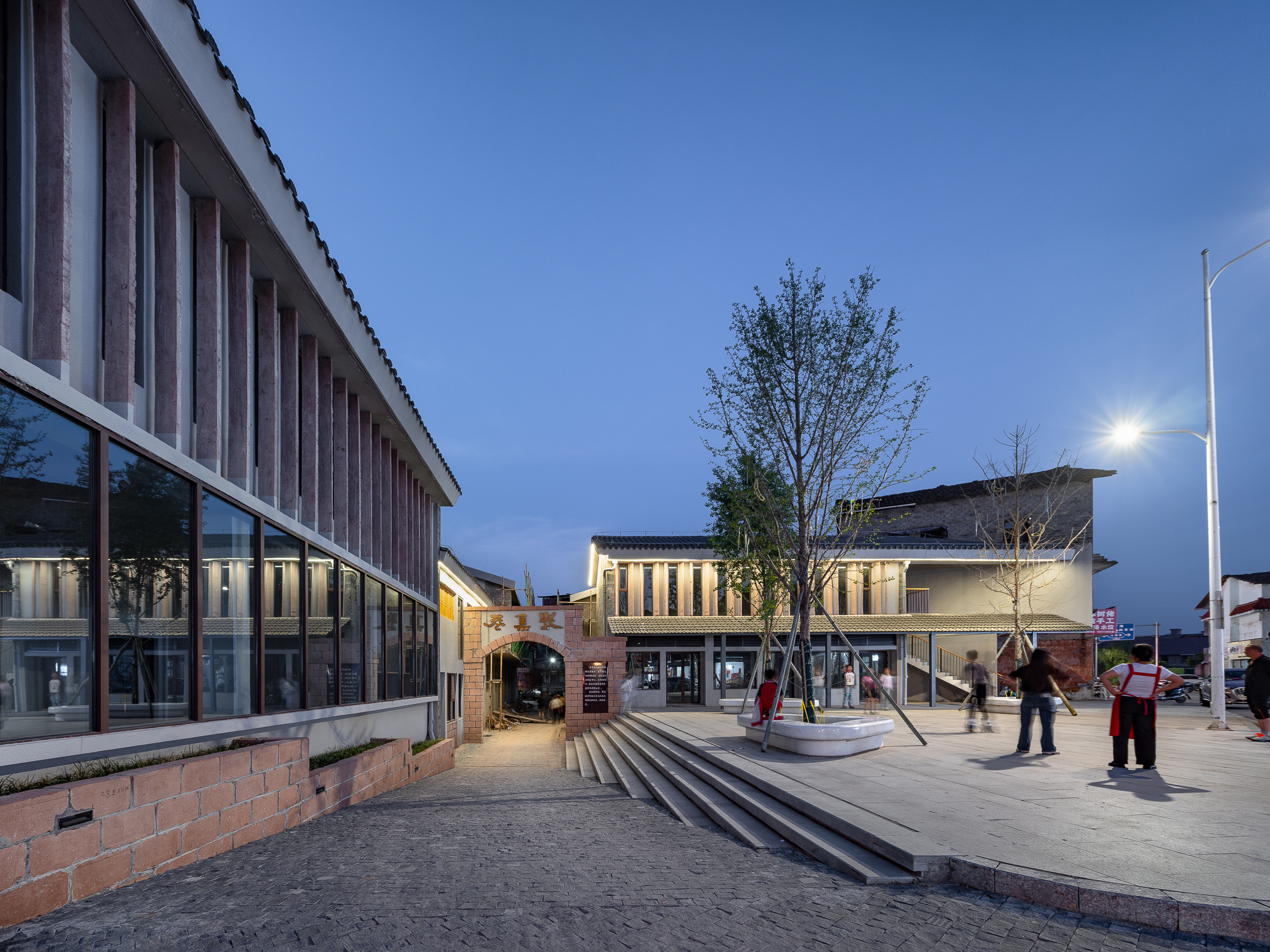 Public plaza with stepped seating and vertical-slatted brick facade under blue evening sky