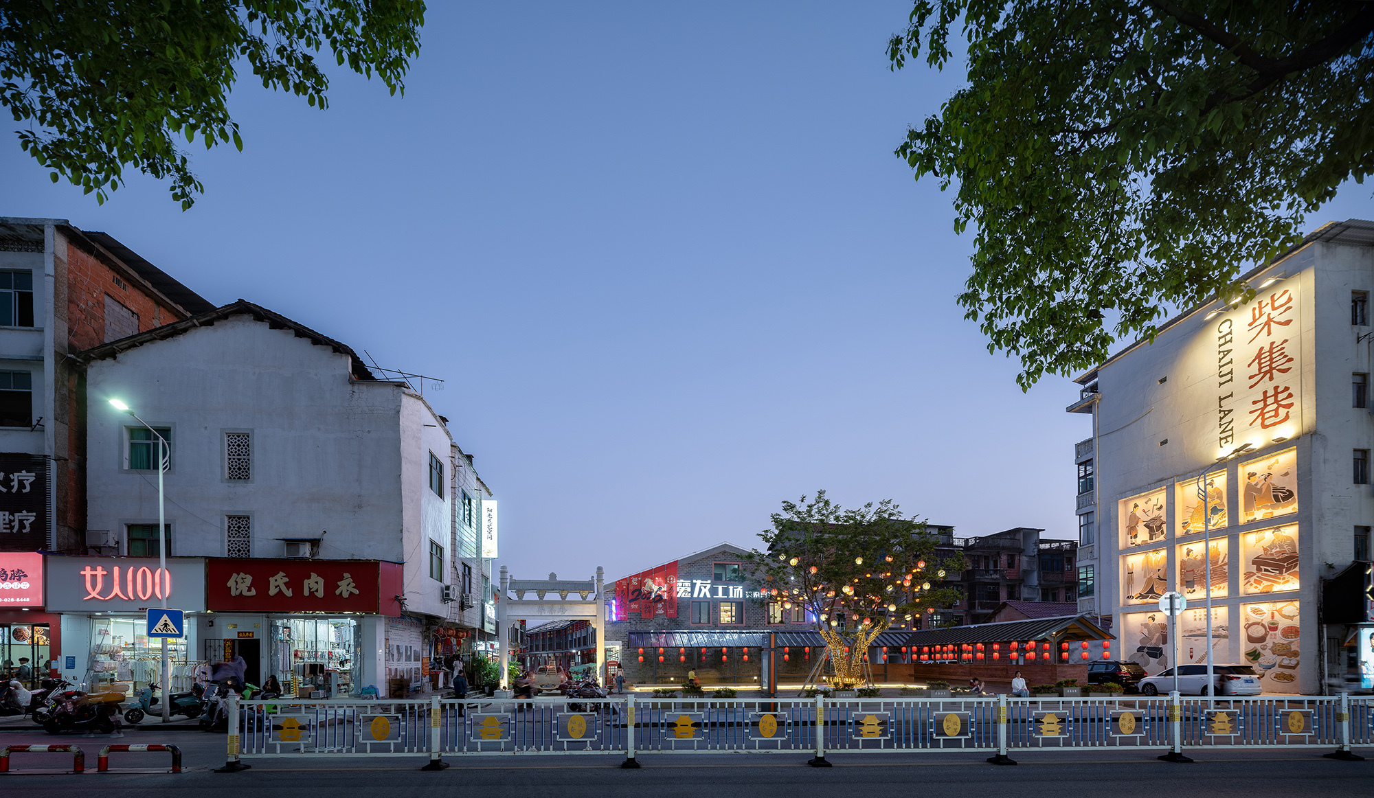 Street view showing mixed commercial buildings with illuminated signage and metal traffic barriers at dusk