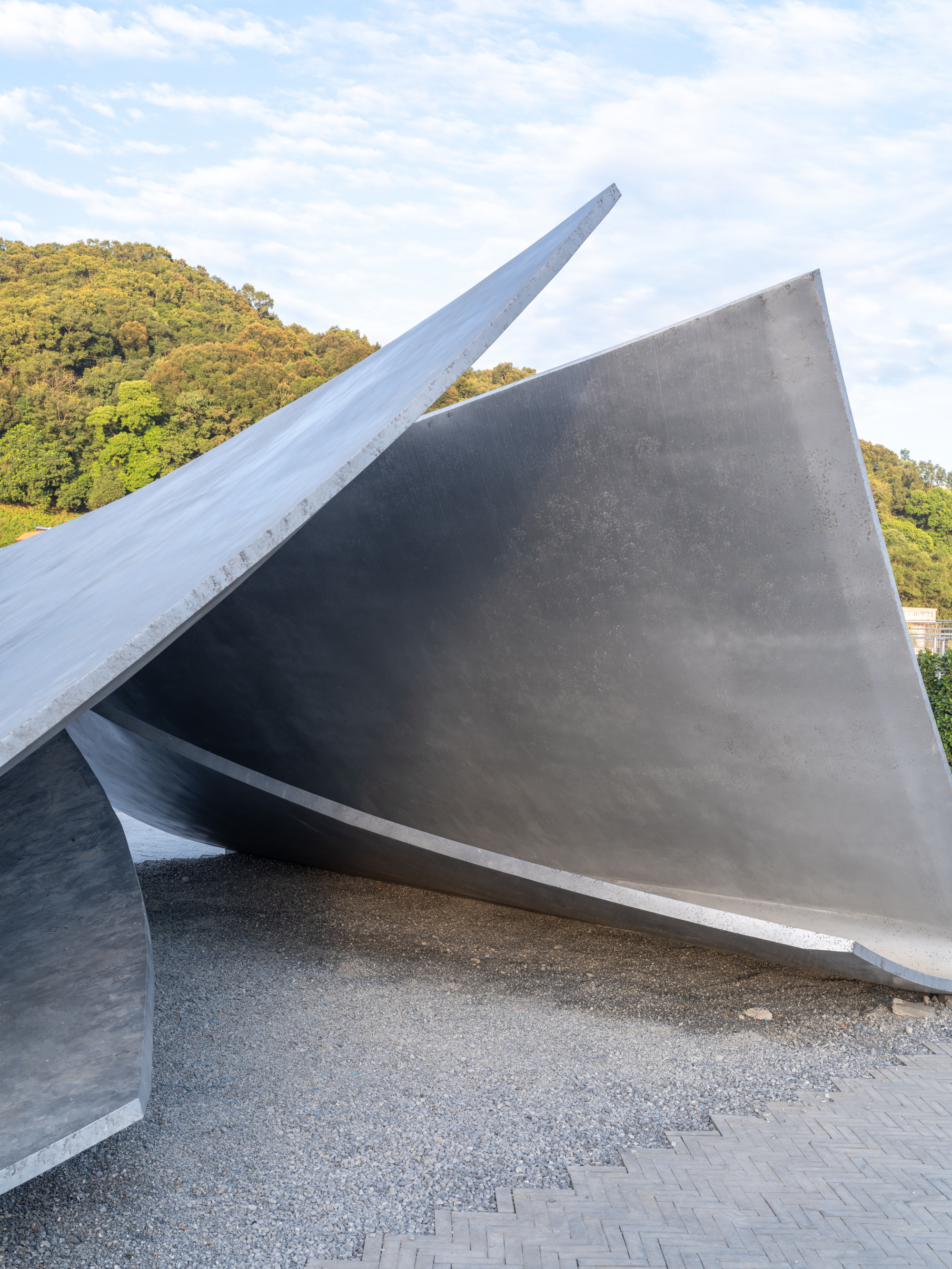 Angled concrete roof planes rising above a gravel plaza with forested hillside in the background