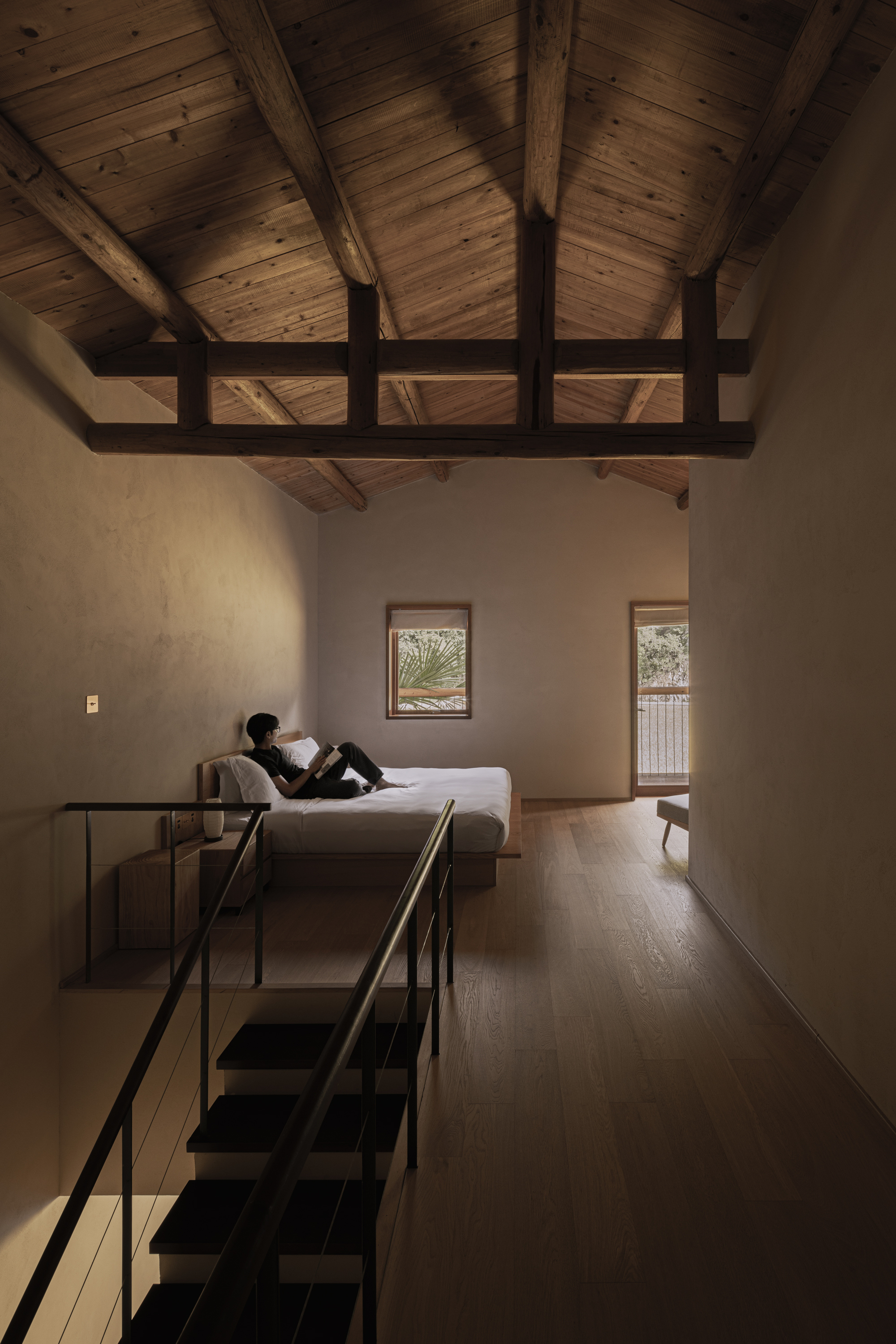 Double-height bedroom with exposed timber roof trusses and a person reading on the daybed