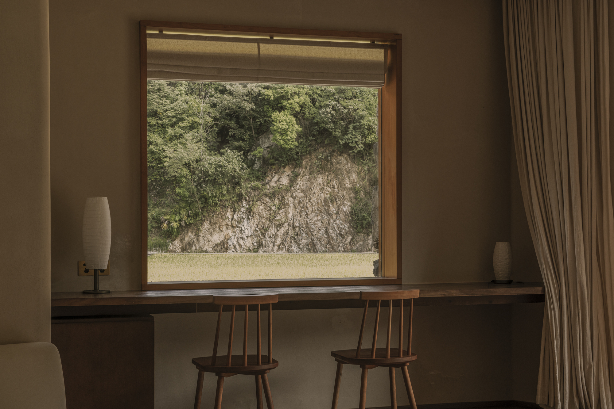 Framed window view of exposed rock outcrop with wooden desk and spindle-back chairs below