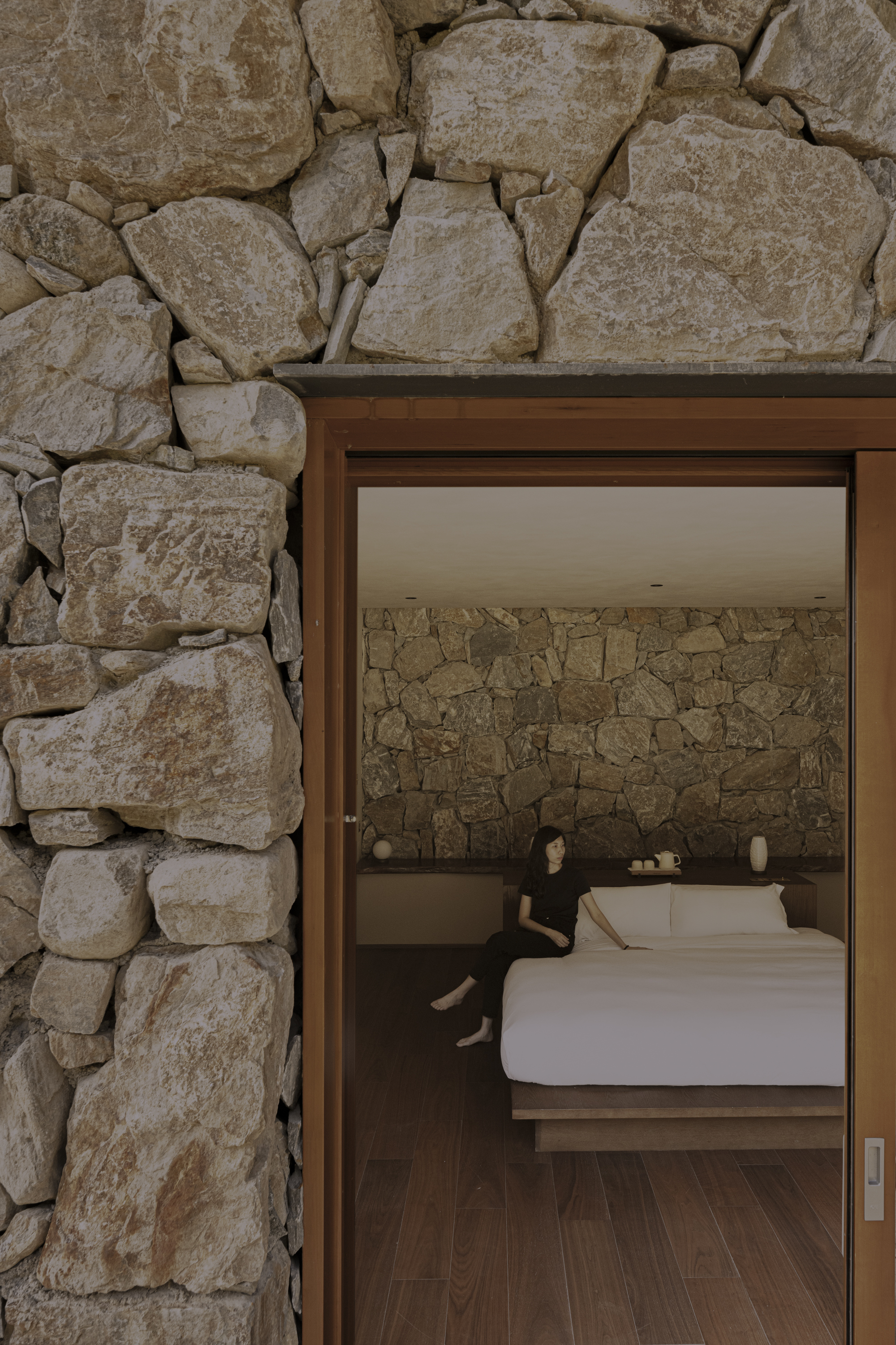 Open doorway through dry-stacked stone wall revealing bedroom with matching stone headboard wall