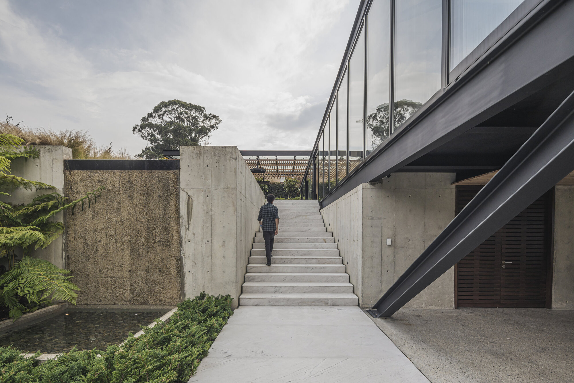 Concrete staircase ascending between board-formed walls with a person climbing under an overcast sky