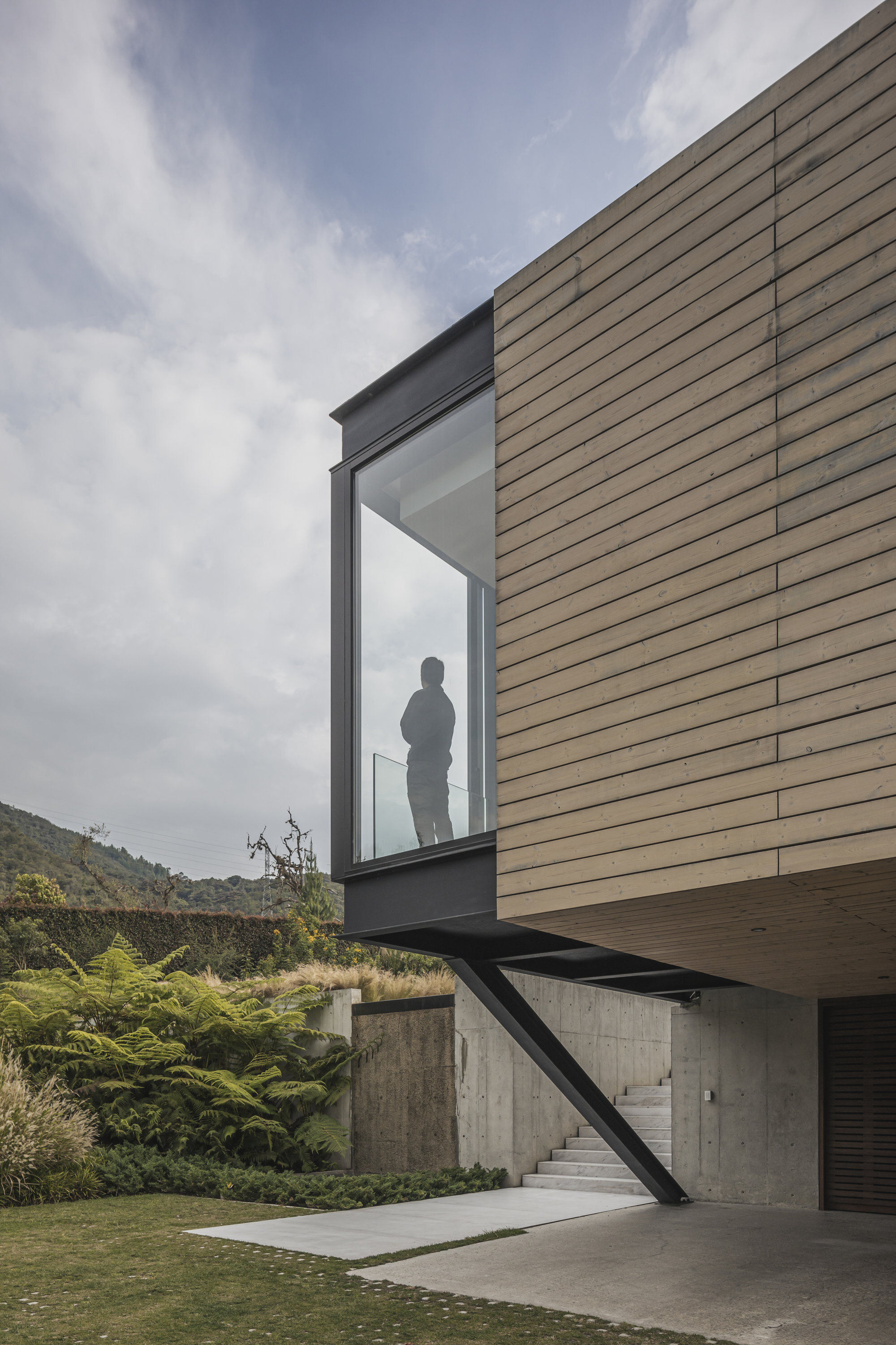 Cantilevered timber-clad bay with a figure silhouetted in the window under cloudy skies