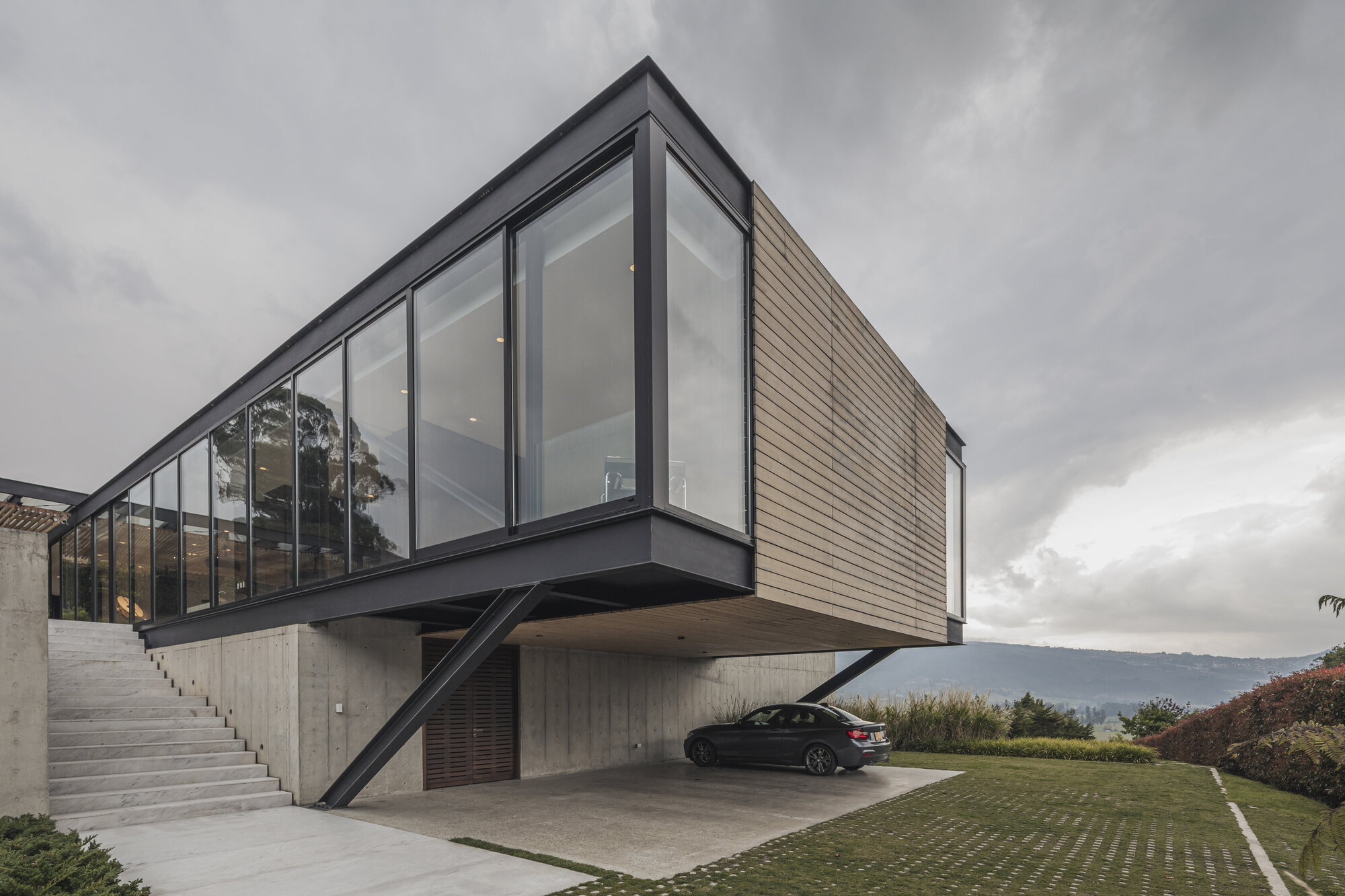 Elevated glass and timber volume cantilevered over a concrete carport with external staircase under overcast skies