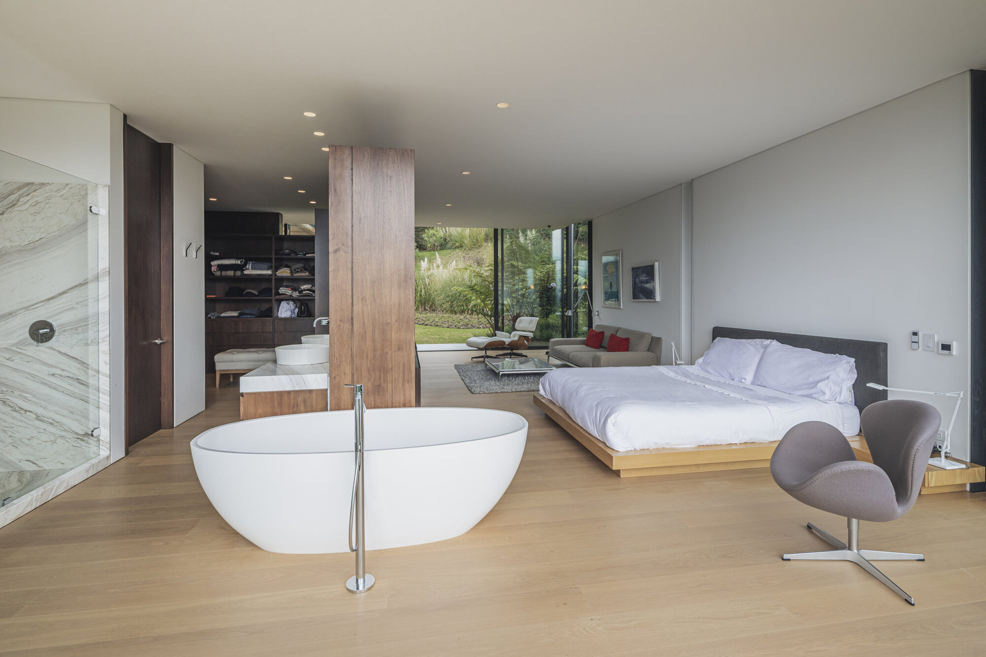 Open bedroom with freestanding white bathtub on timber flooring and glazed walls facing vegetation