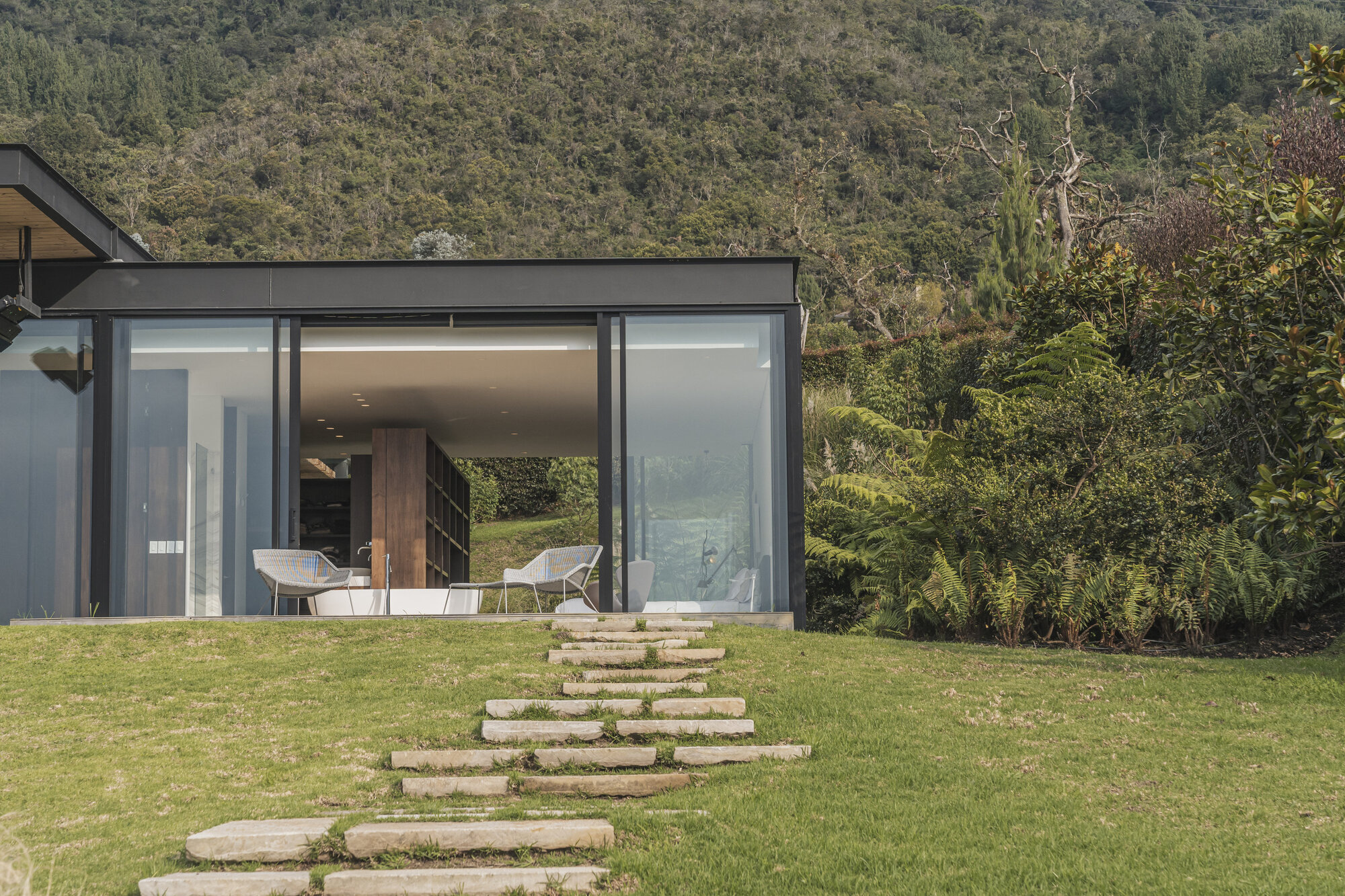 Glass-walled pavilion with stone stepping path crossing lawn toward forested hillside
