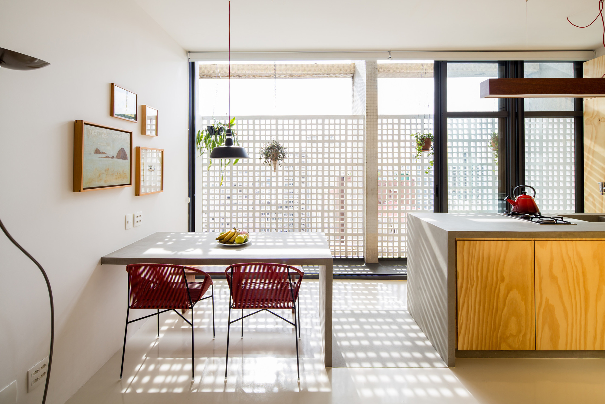 Dining table and red woven chairs before a perforated concrete screen, with sunlight casting dotted shadows across the floor