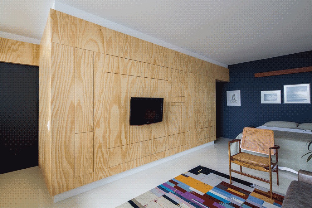 Plywood wall unit with a wall-mounted television beside a navy bedroom wall, cane armchair and striped rug in daylight