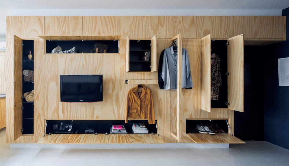 Plywood storage wall with open doors and fold-down shelves showing a hanging jacket, shirt, shoes, books and rolled towels
