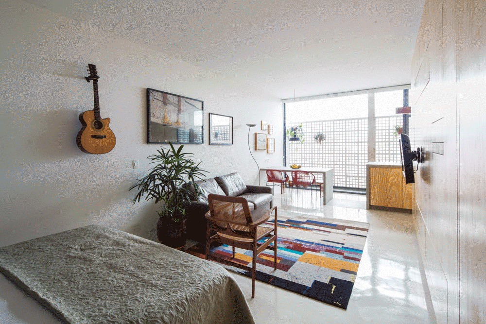 Open living space seen from the bed, with wall-mounted guitar, leather sofa, potted plant and colorful rug leading to a screened window