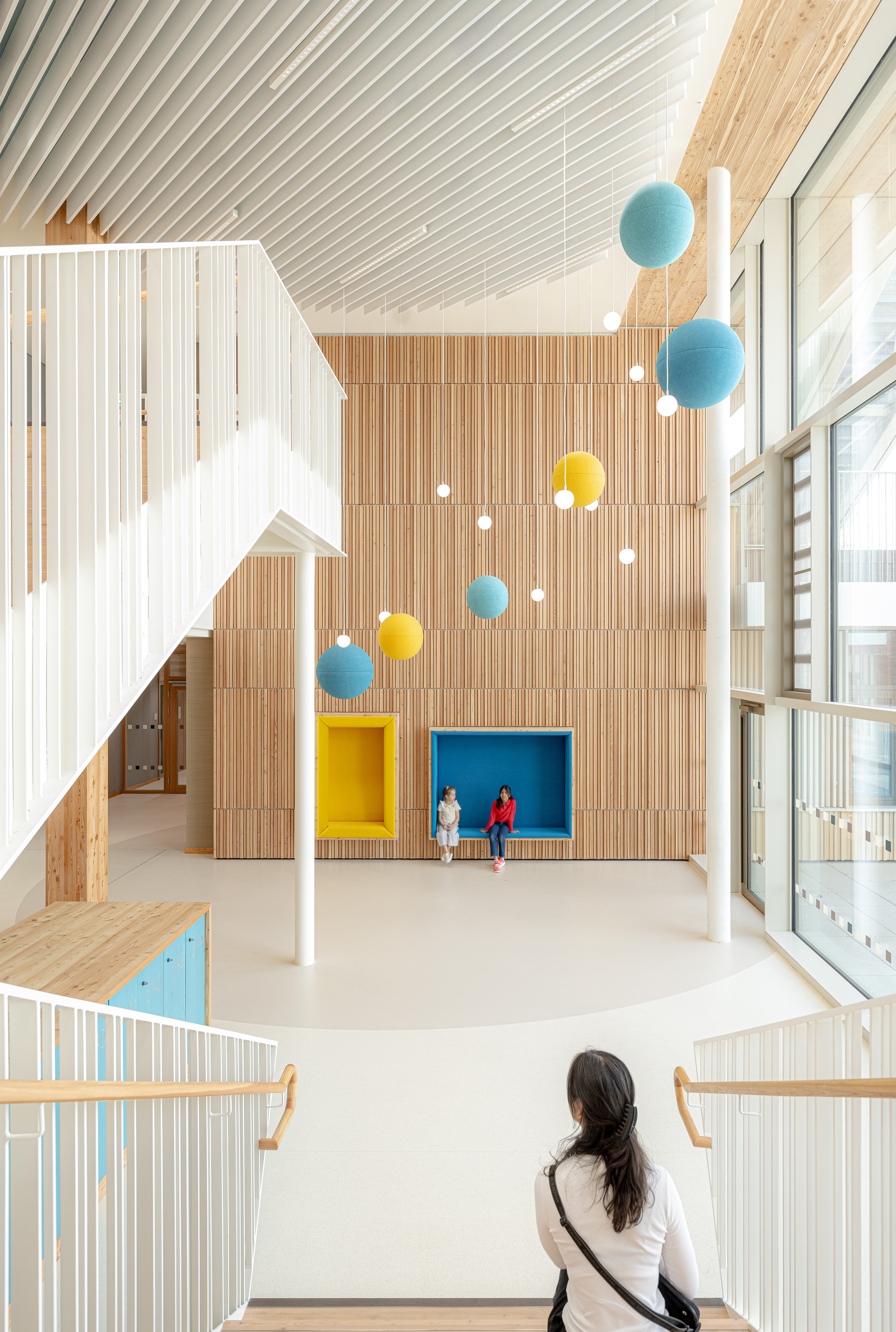 Double-height atrium with vertical timber cladding, suspended sphere pendants, and a figure on the upper balcony
