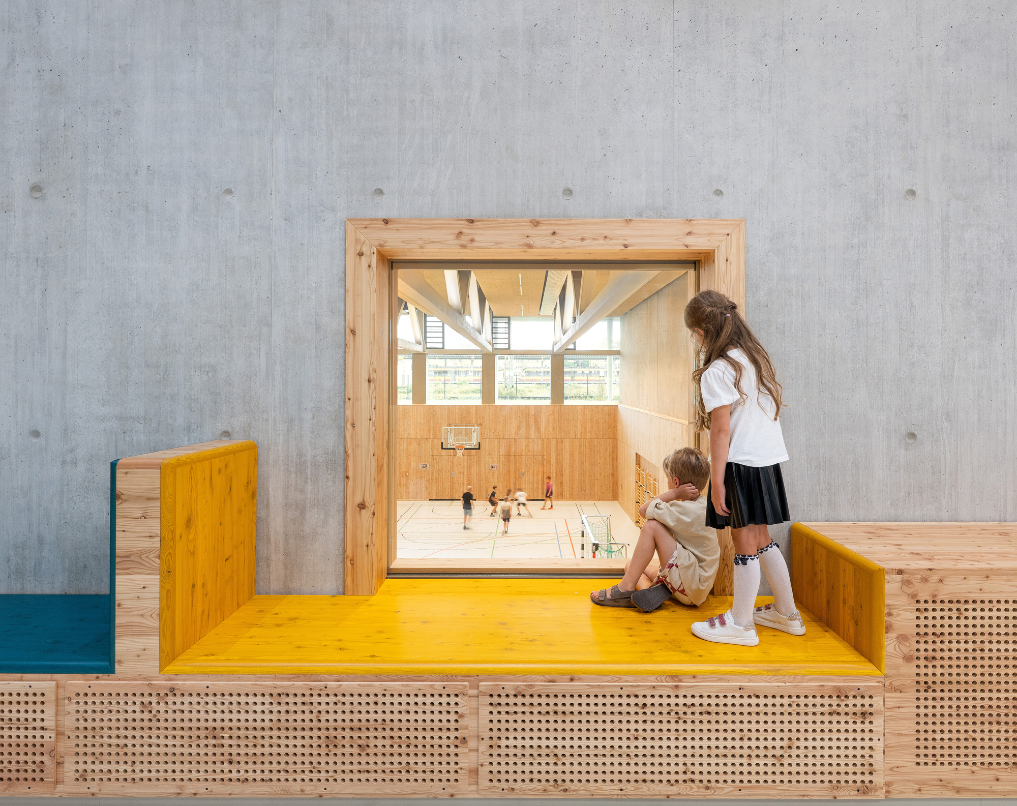 Timber-framed observation window set into a board-formed concrete wall, with two children looking through