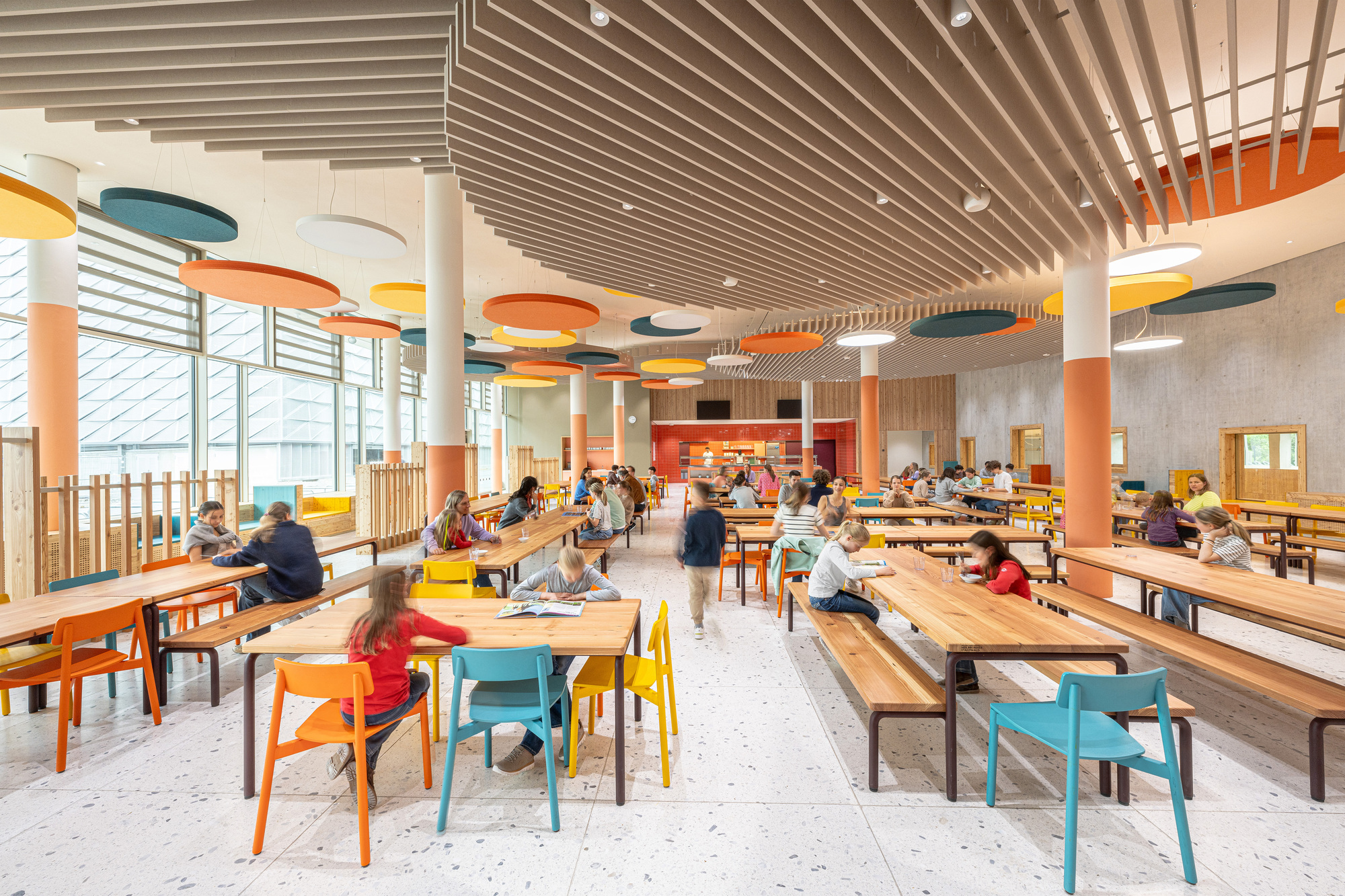 Dining hall with suspended circular acoustic panels, timber benches, and students seated at tables