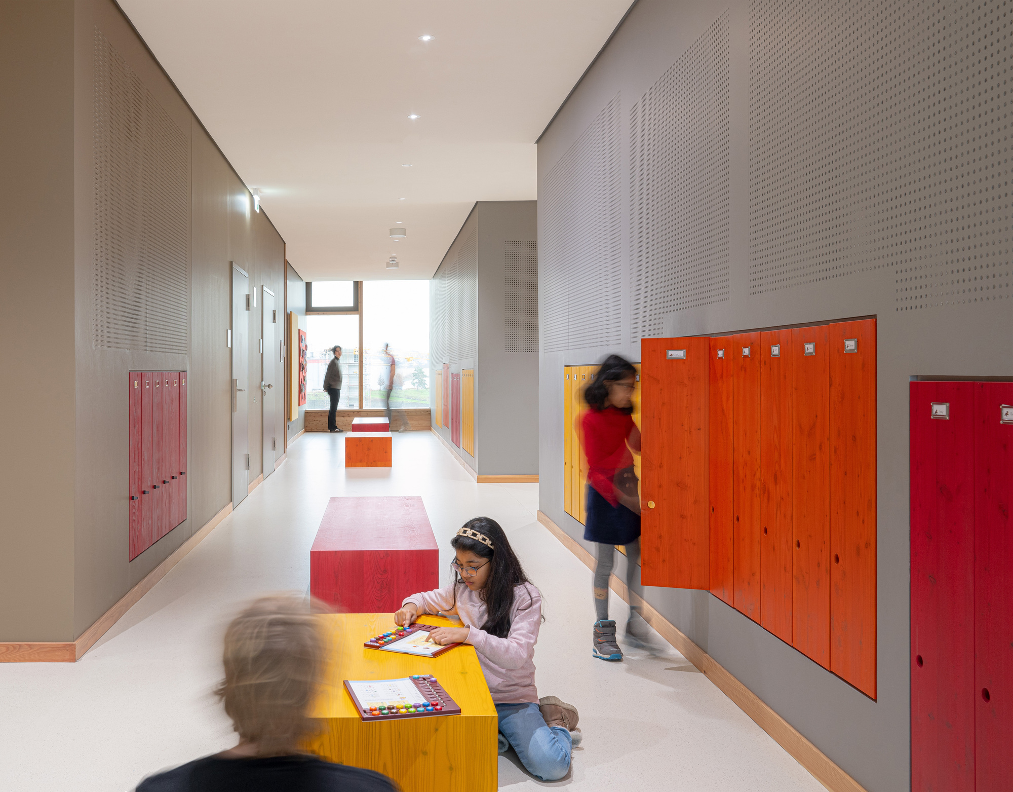White corridor with perforated metal walls, colorful lockers and children sitting on modular seating blocks
