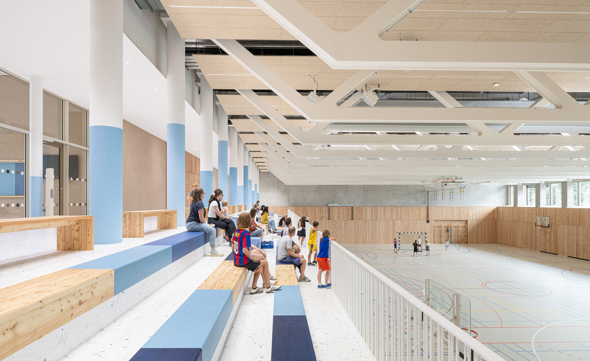 Indoor sports hall with tiered bleacher seating, plywood ceiling panels and children on the court floor