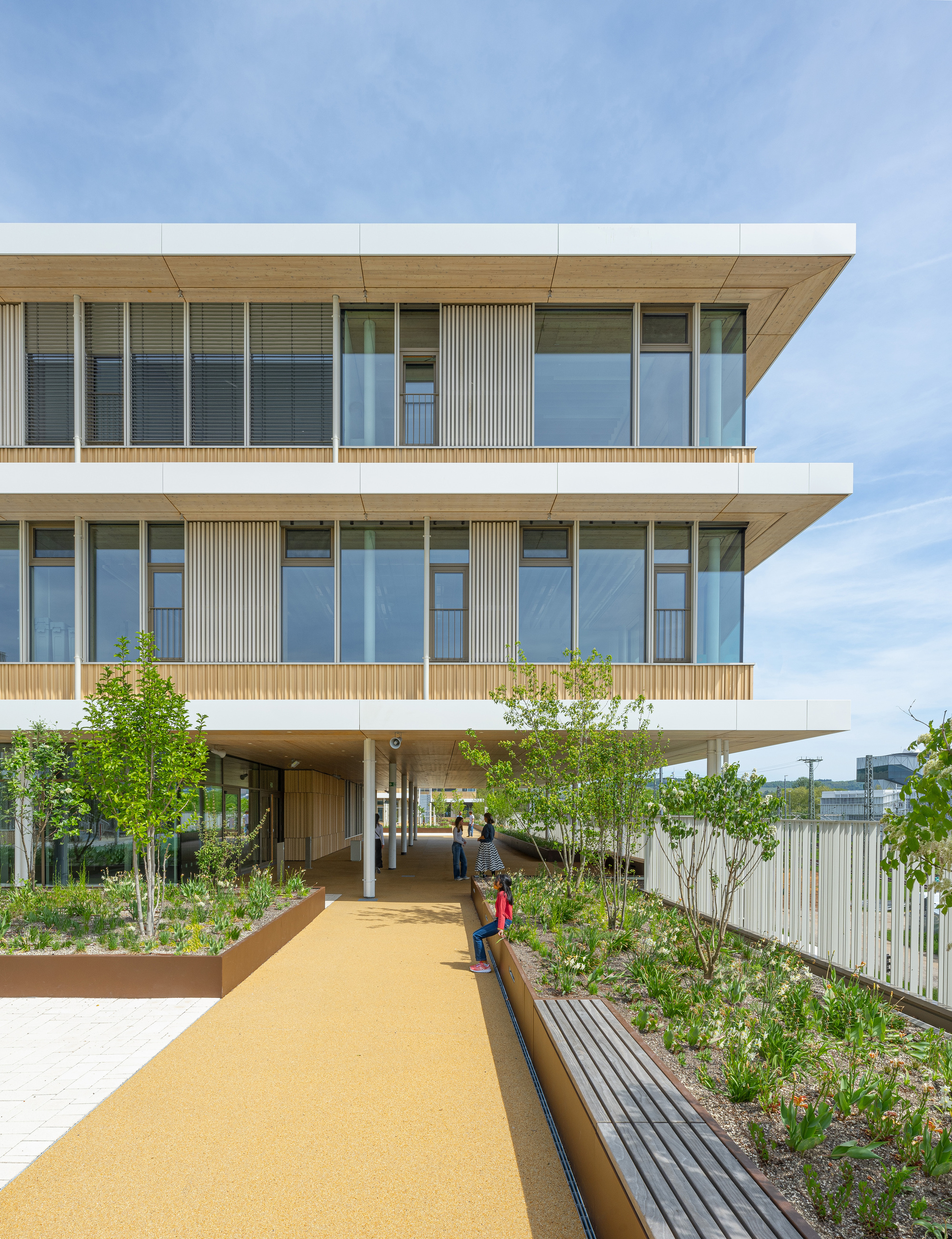 Raised walkway with timber benches and planted beds leading to a piloti ground floor under a clear sky