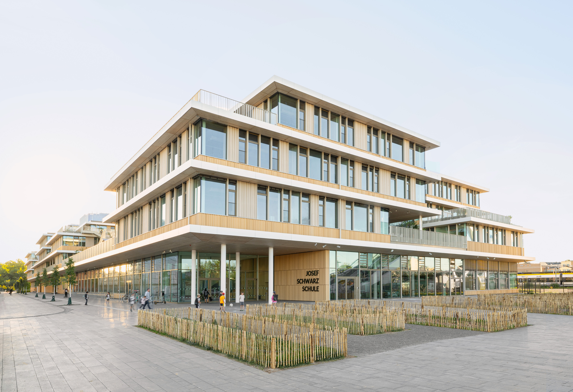 Corner view of the timber and glass facade with stacked floor plates and planted beds at the plaza